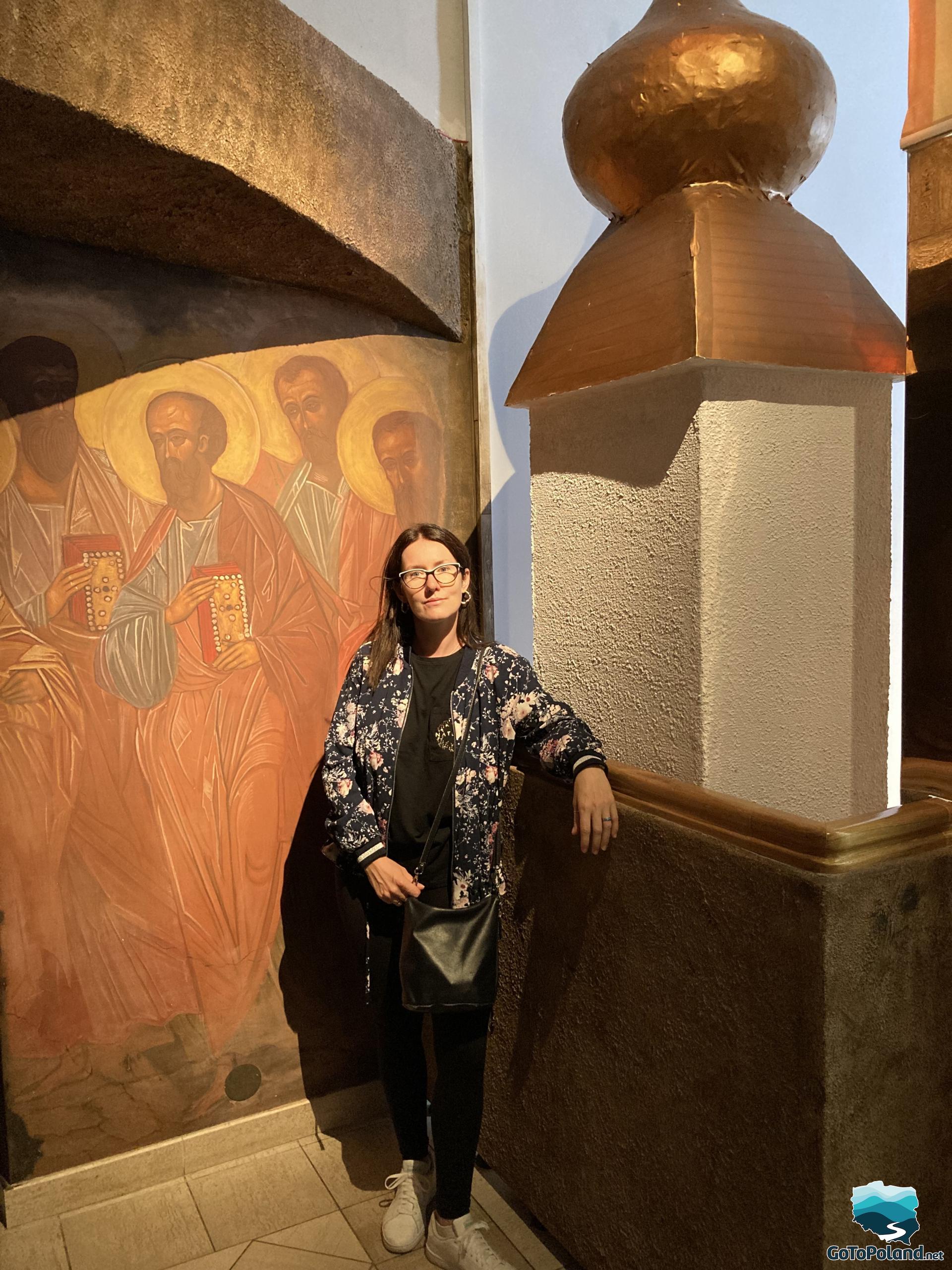 a woman stands in front of a wall on which images of saints are painted