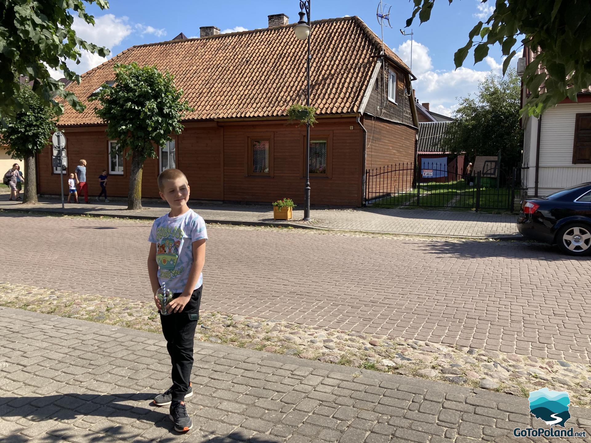 the boy is standing by the street, behind him a brown wooden house