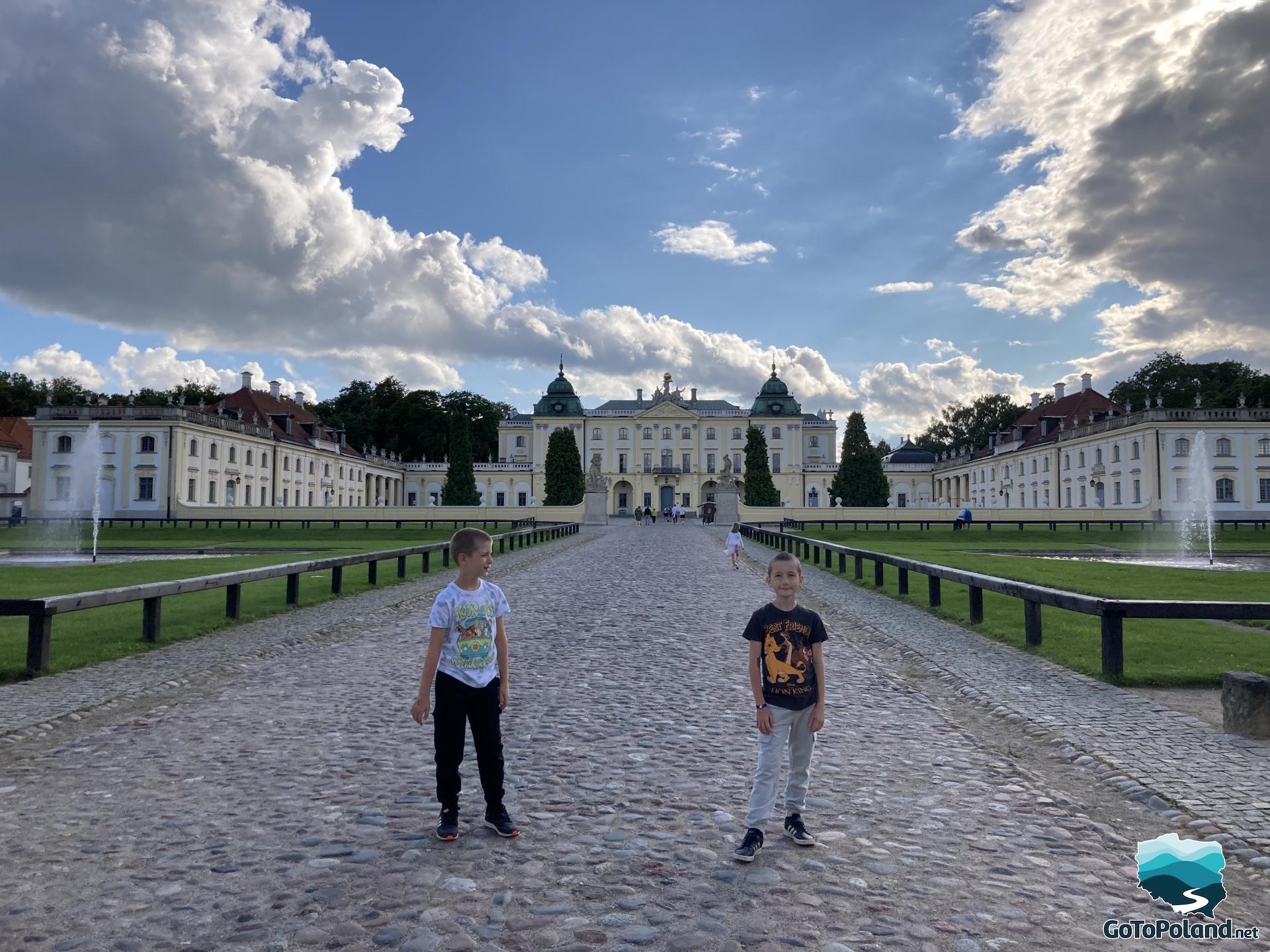 two boys are standing on the path leading to the palace which is in the background. The facade is yellow and white. In front of the palace, on both sides of it, there are two fountains.
