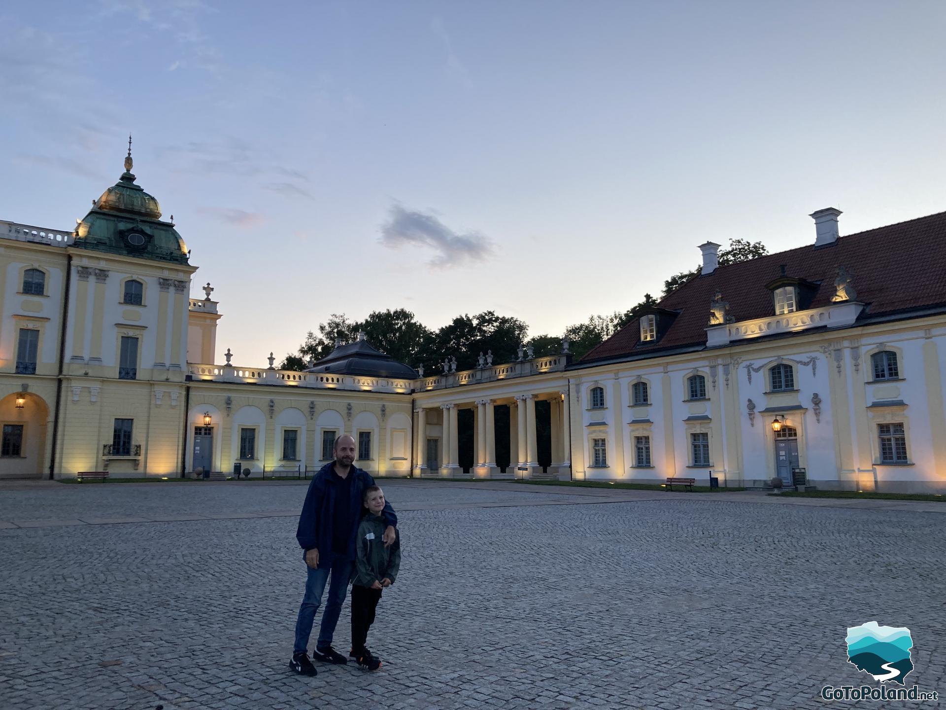 a man and a boy are standing in front of the castle, the palace is illuminated because it is already evening