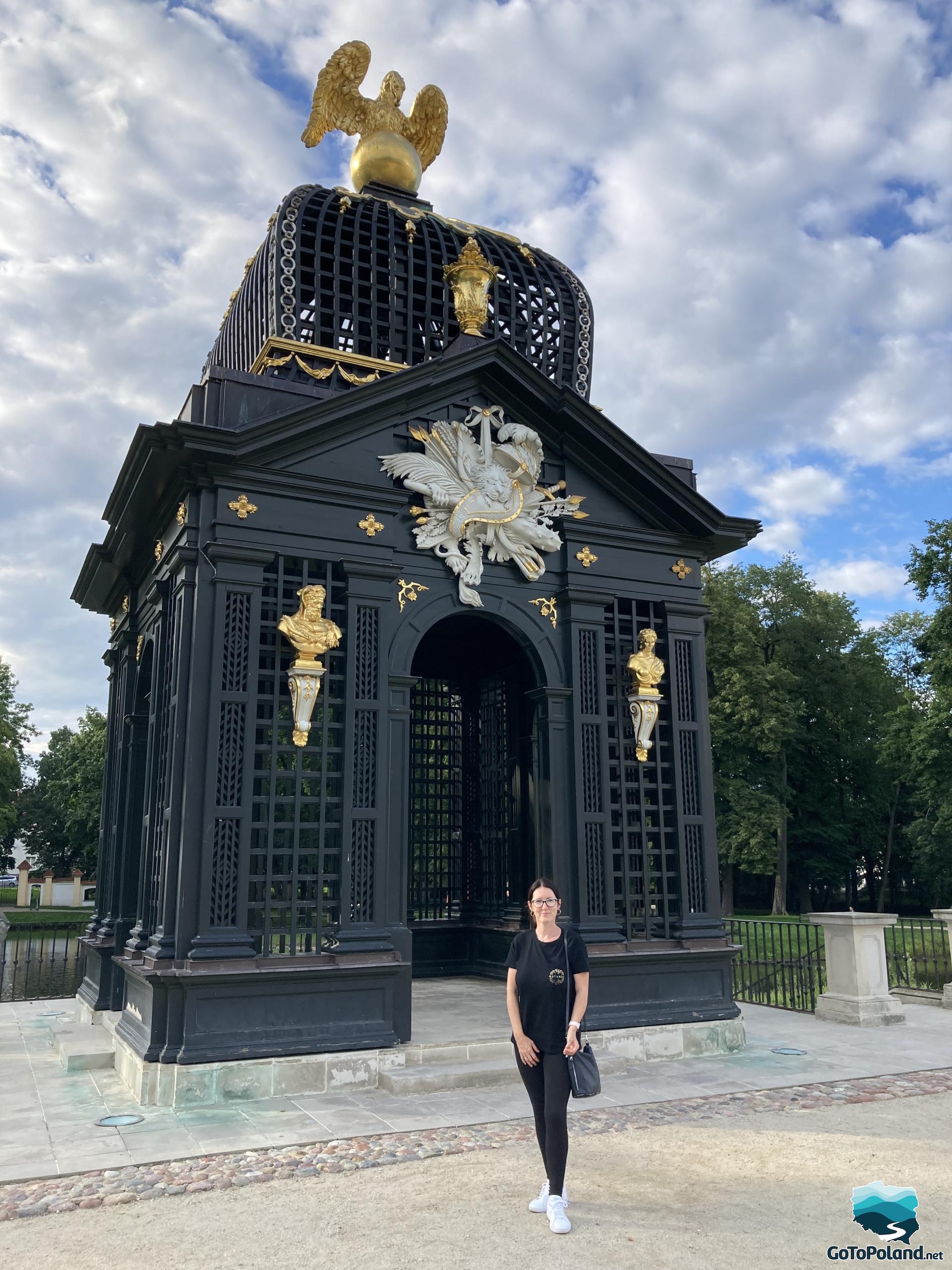 a black great gate with a gold statue of a bird on top, on the gate there are gold ornaments, a woman is standing in front of it 