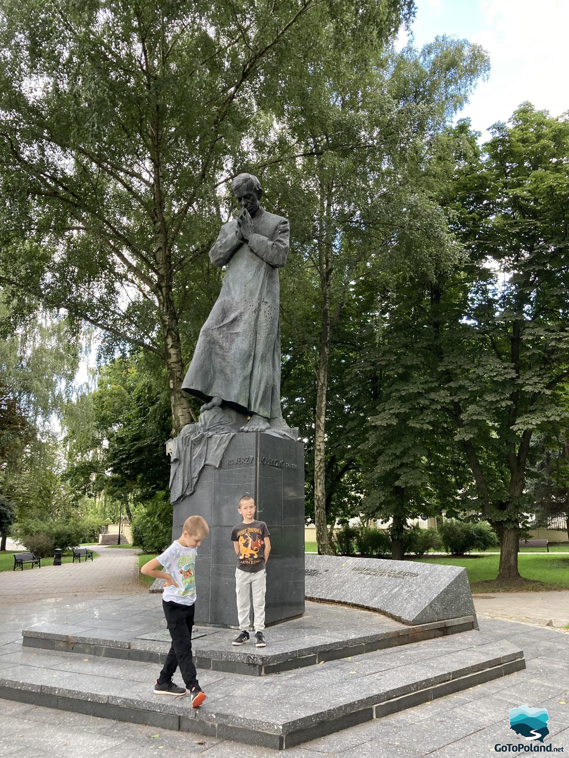 a statue of priest Jerzy Popieluszko on a pedestal, two boys are by the statue