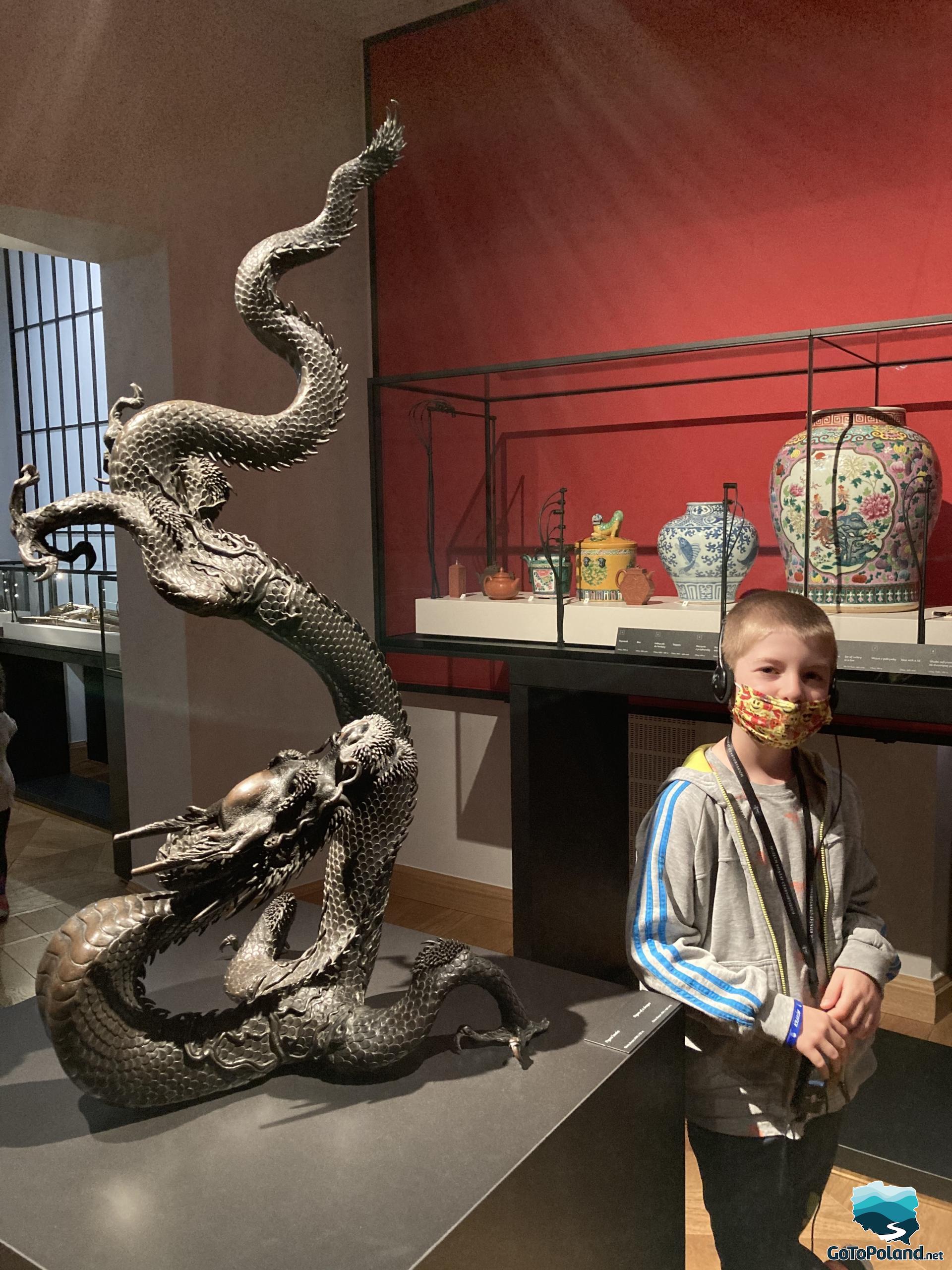 A boy is standing by the figure of a snake. Behind him there are some vases 