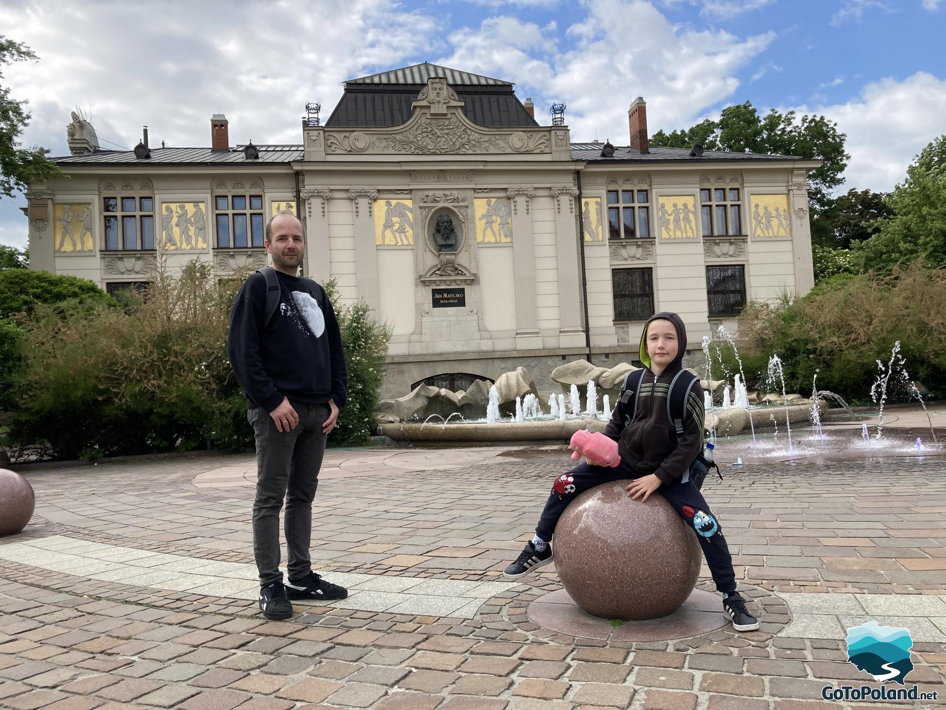 a man is standing, next to him a boy is sitting on a ball made of stone, behind them a fountain and a decorative building with a bust of Jan Matejko