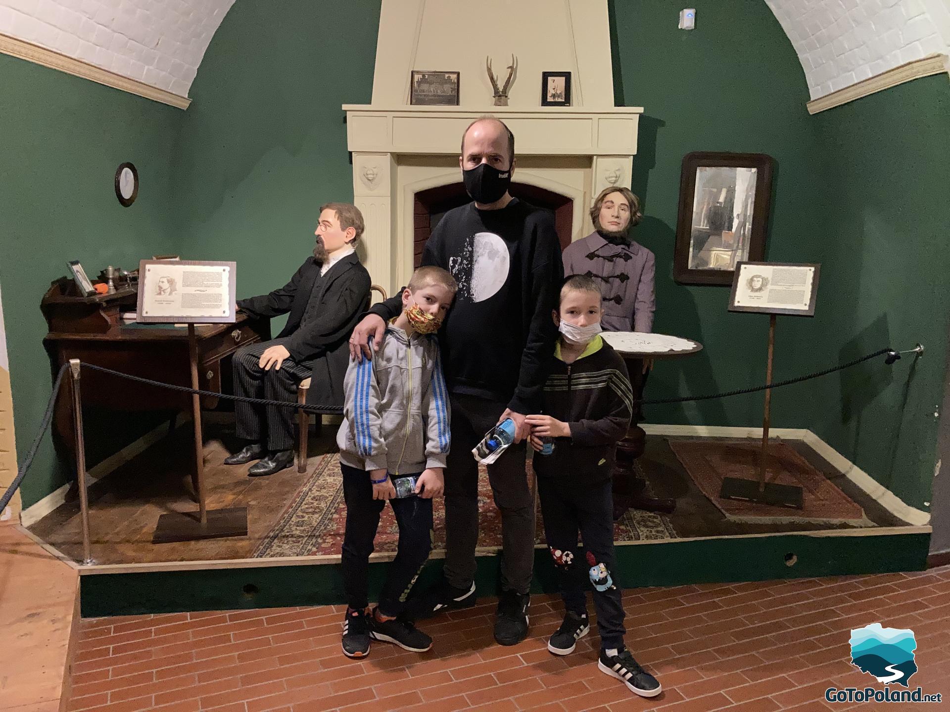 A man and two kids are standing in front of two wax figures of polish writers.