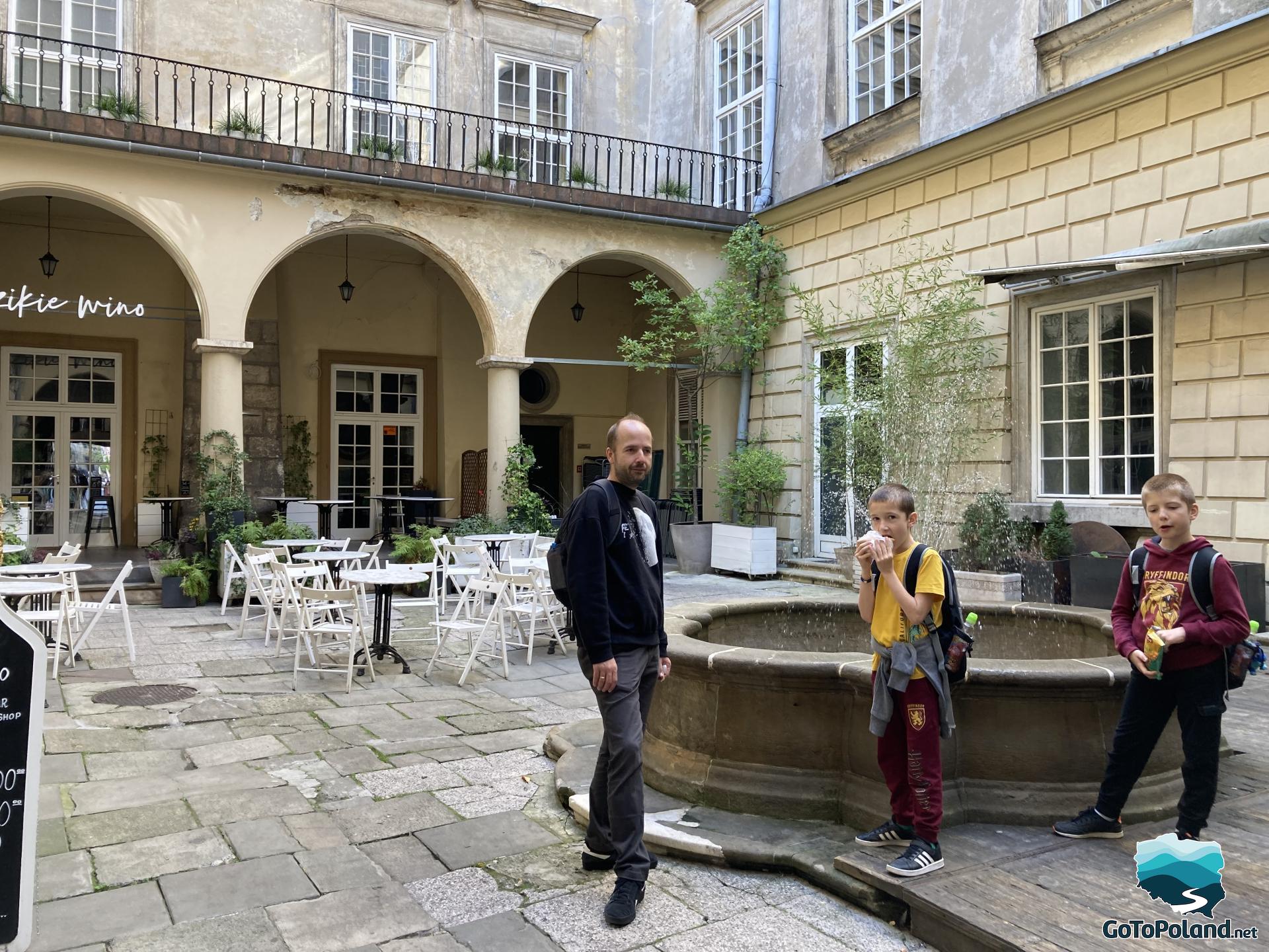 a man with two children is standing by a stone fountain, they are standing in a square between tenement houses, in the background white chairs and tables belonging to a restaurant