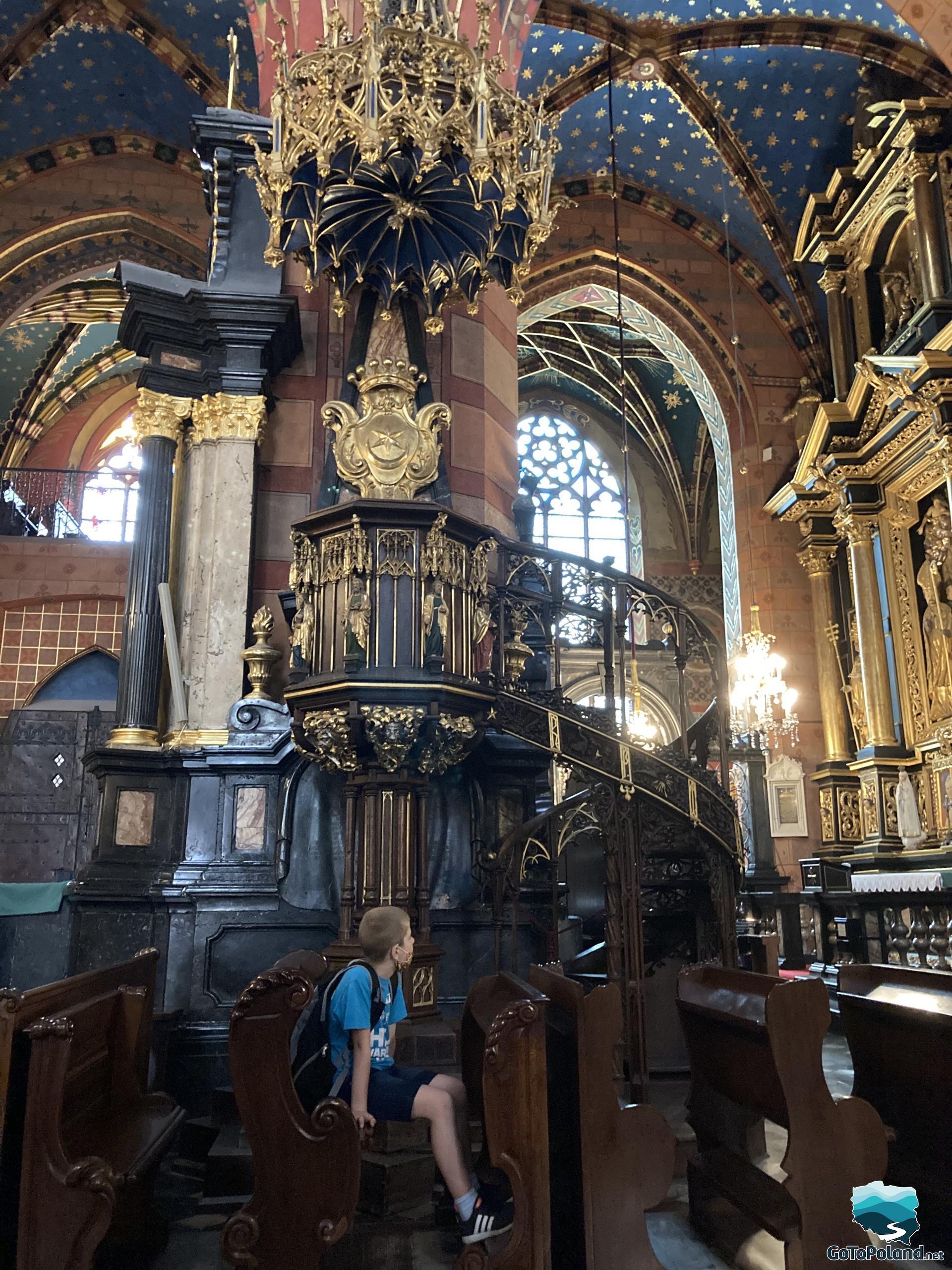 a boy is sitting in a pew, above him, there is a black and gold pulpit, ceiling of the church is blue