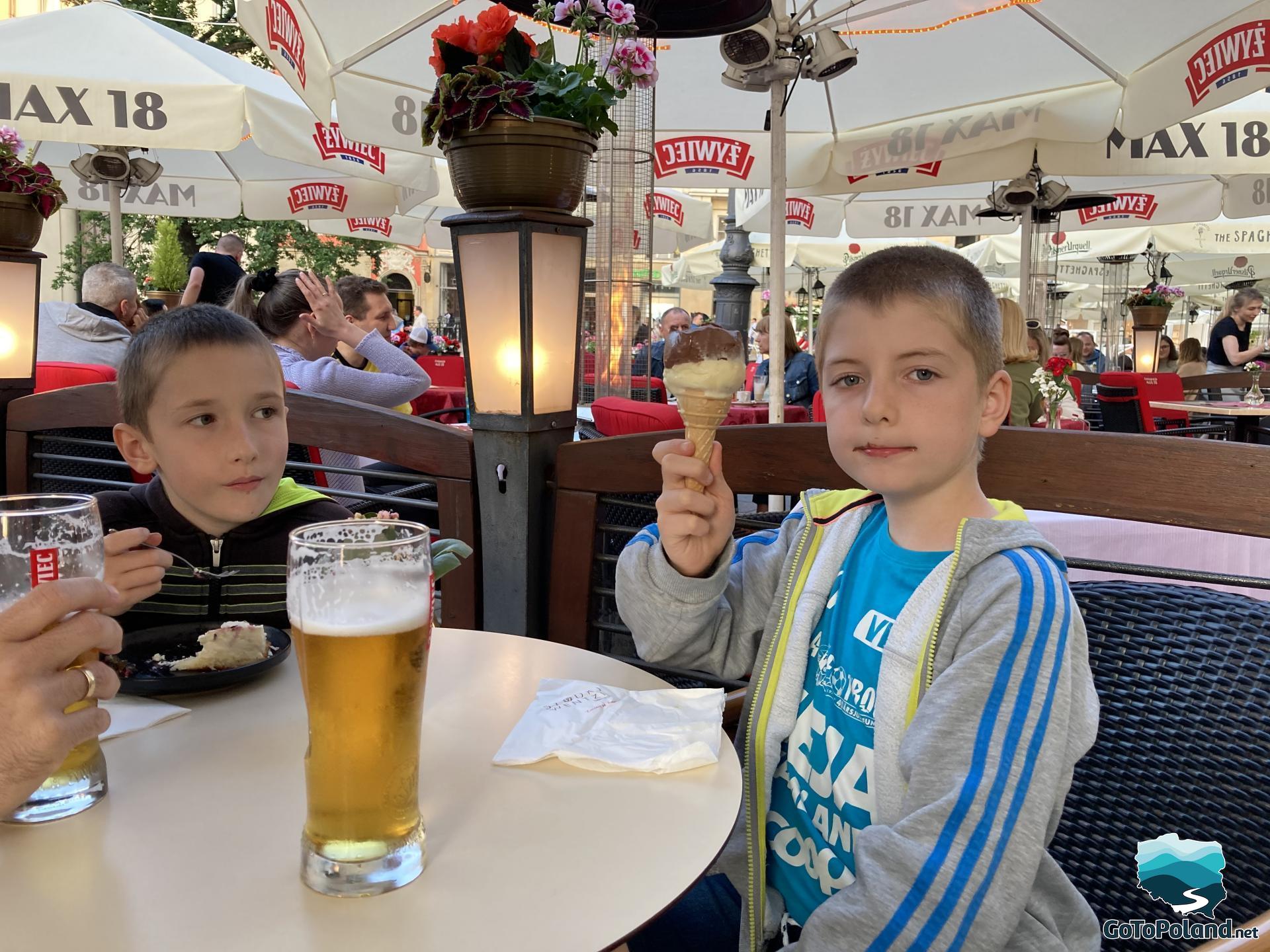 a family is sitting in a outside restaurant, a glass of beer is on the table, one boy is eating an ice-cream, the second boy is eating a cake