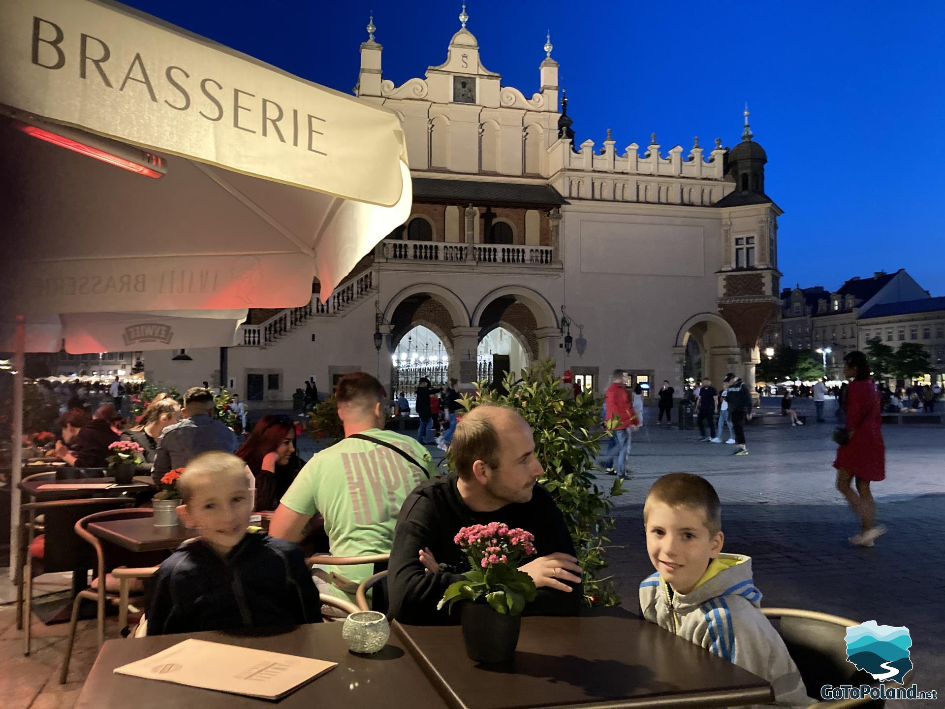 its evening, the family is sitting at a table in an open-air restaurant, the Cloth Hall behind them