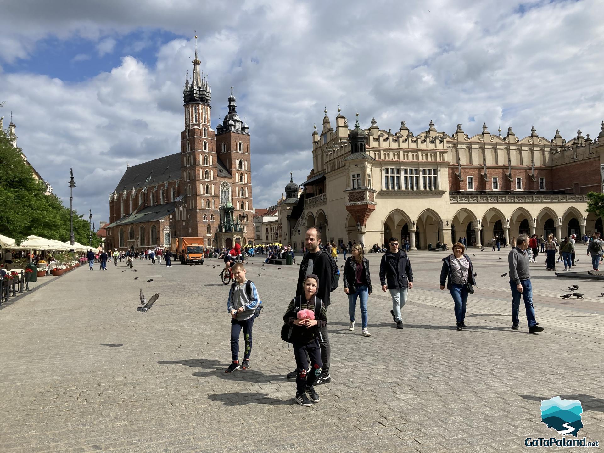 People are on the main square, you can see also pigeons and two buildings: one is a high church in Gothic style, the second building is quite bright with small towers on top