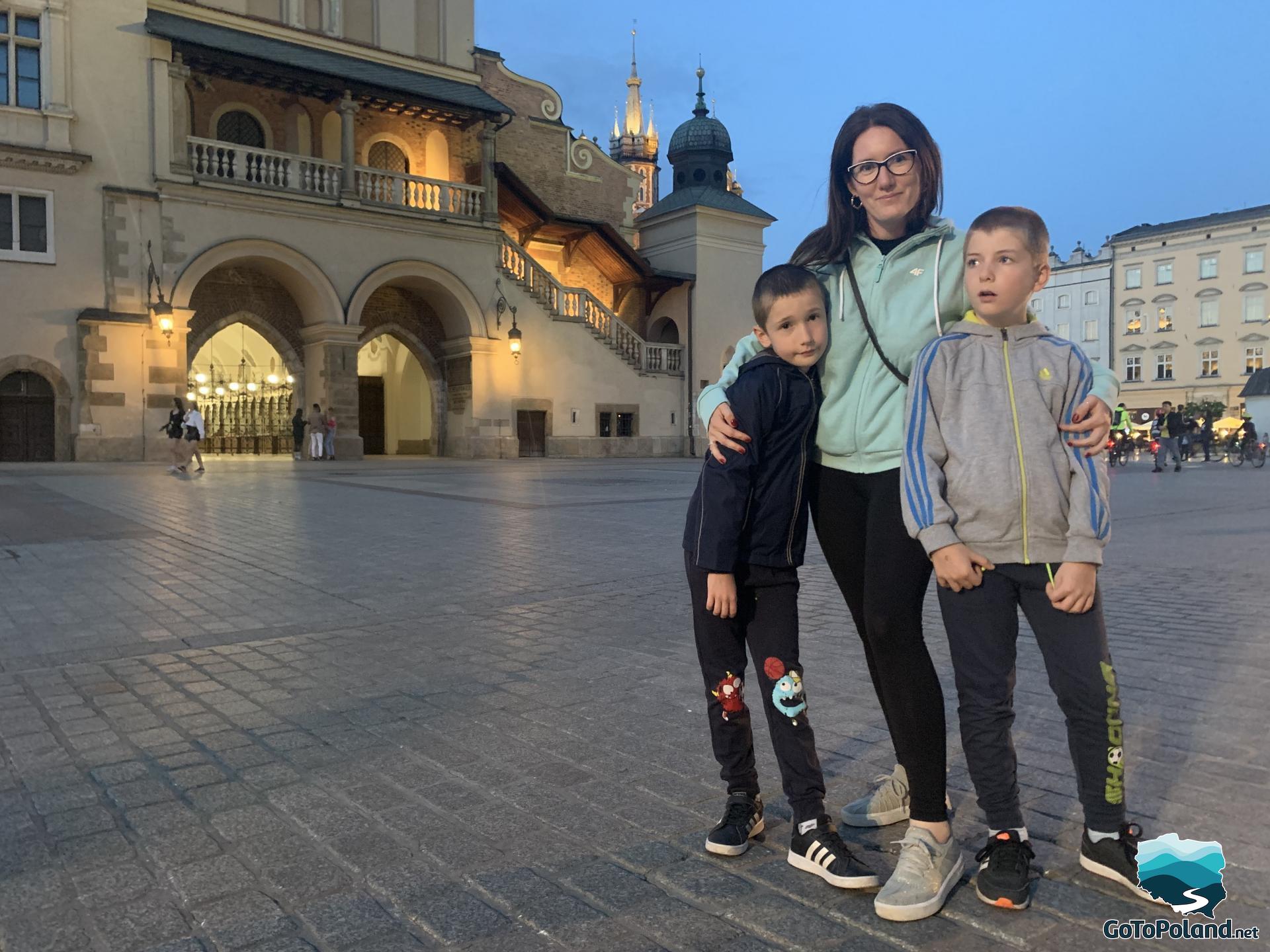 it is evening, a woman and two kids are standing in front of the Cloth Hall 