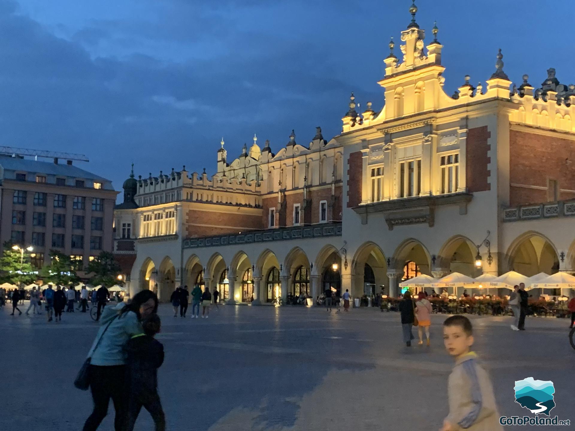People are on the Main Square, the historical building the Cloth Hall is illuminated, its evening 