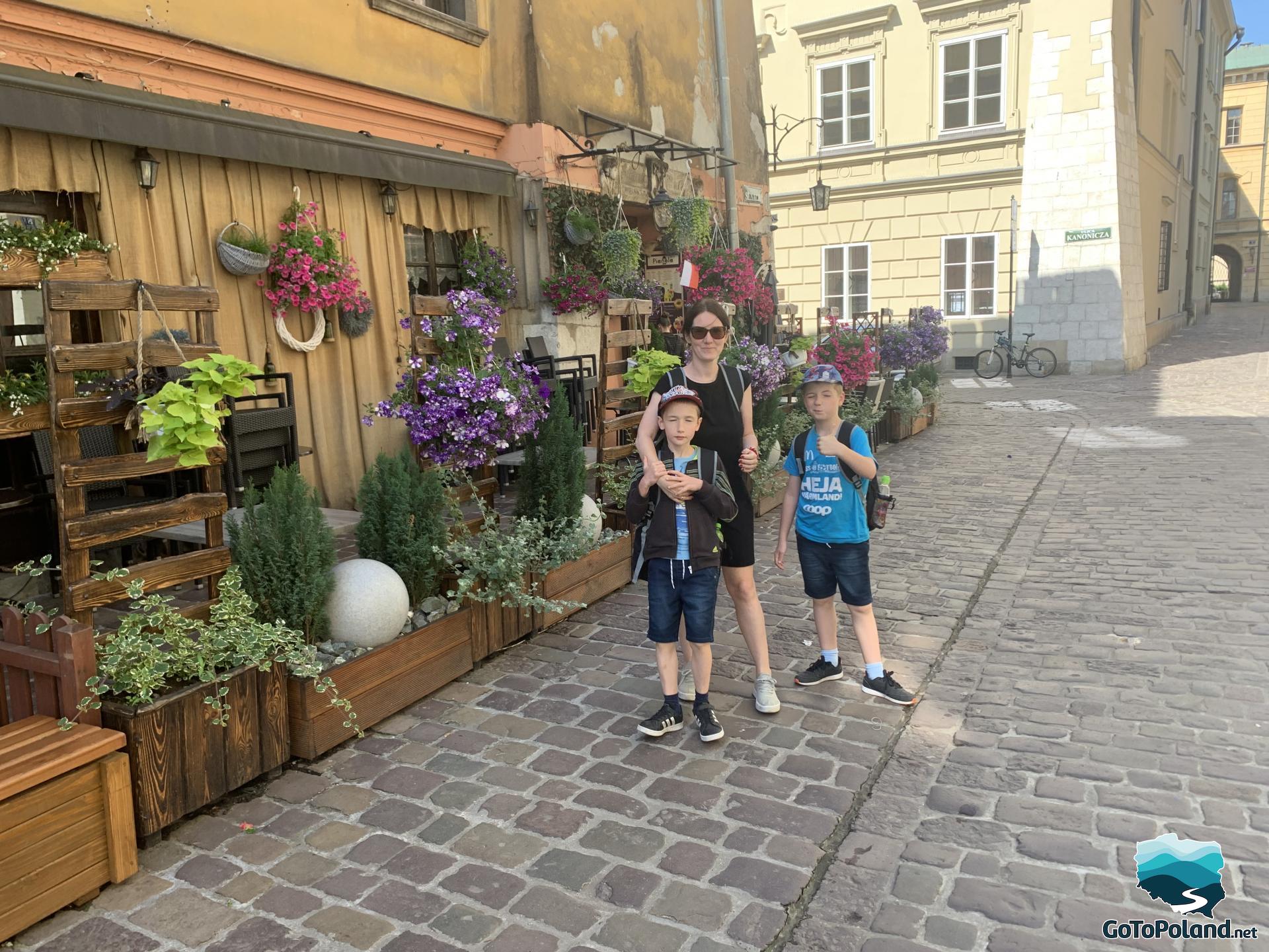 cobbled alley, on the left lots of flowers, two boys and a woman are standing next to the flowers
