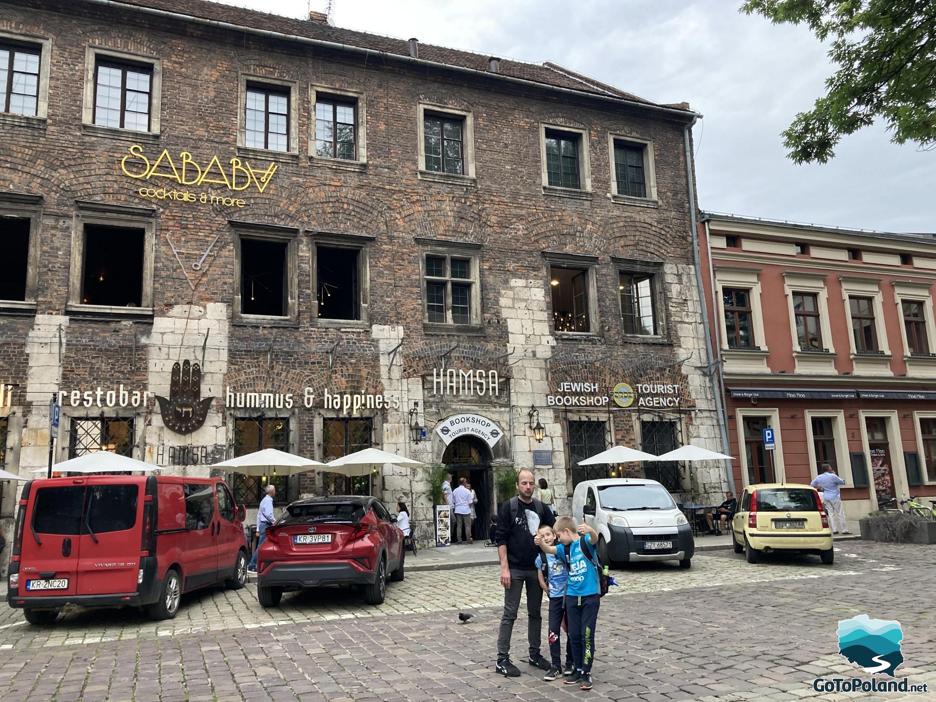a man with two children are standing front on a restaurant, the restaurant is in a historical building, behind man and two boys are 4 modern cars