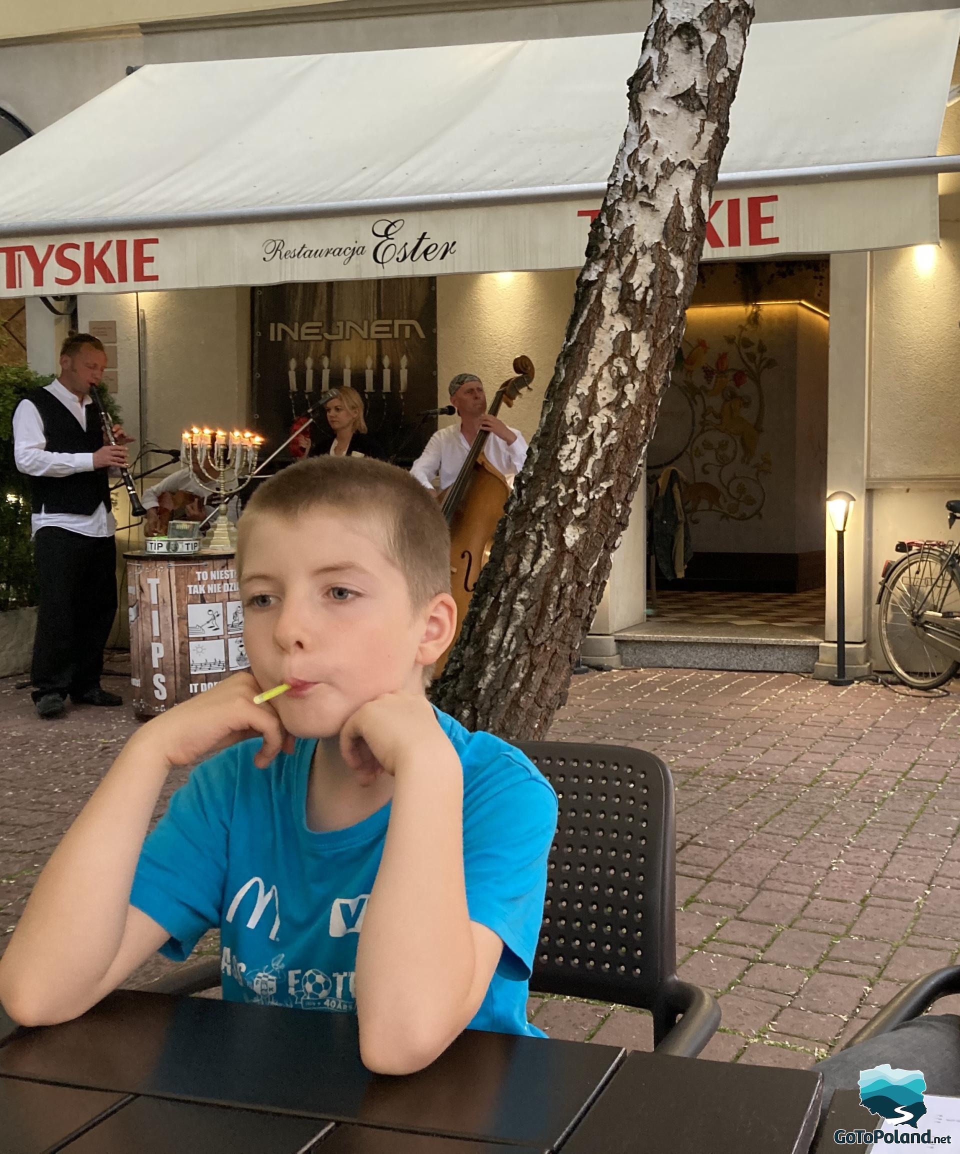 a boy is sitting by the table in an open-air restaurant, behind him there is a band playing Jewish music