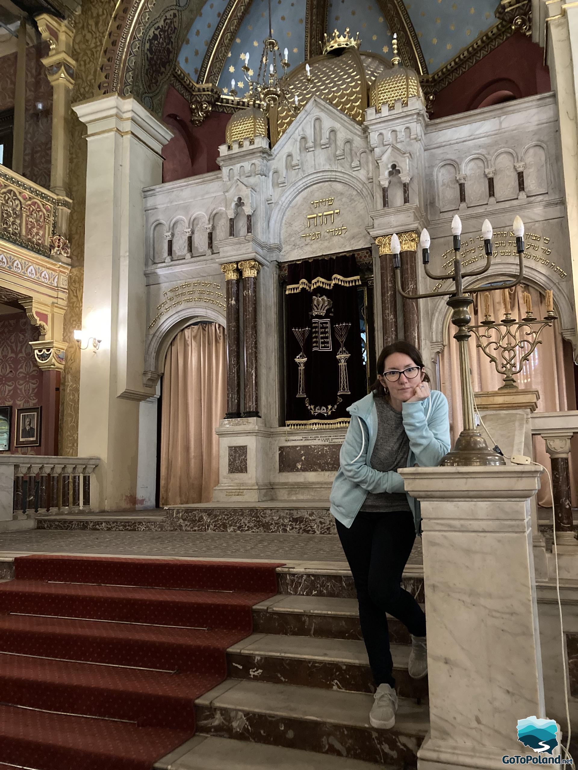 a woman is standing in a synagogue, behind her there is an altar cabinet used to store the Torah scrolls