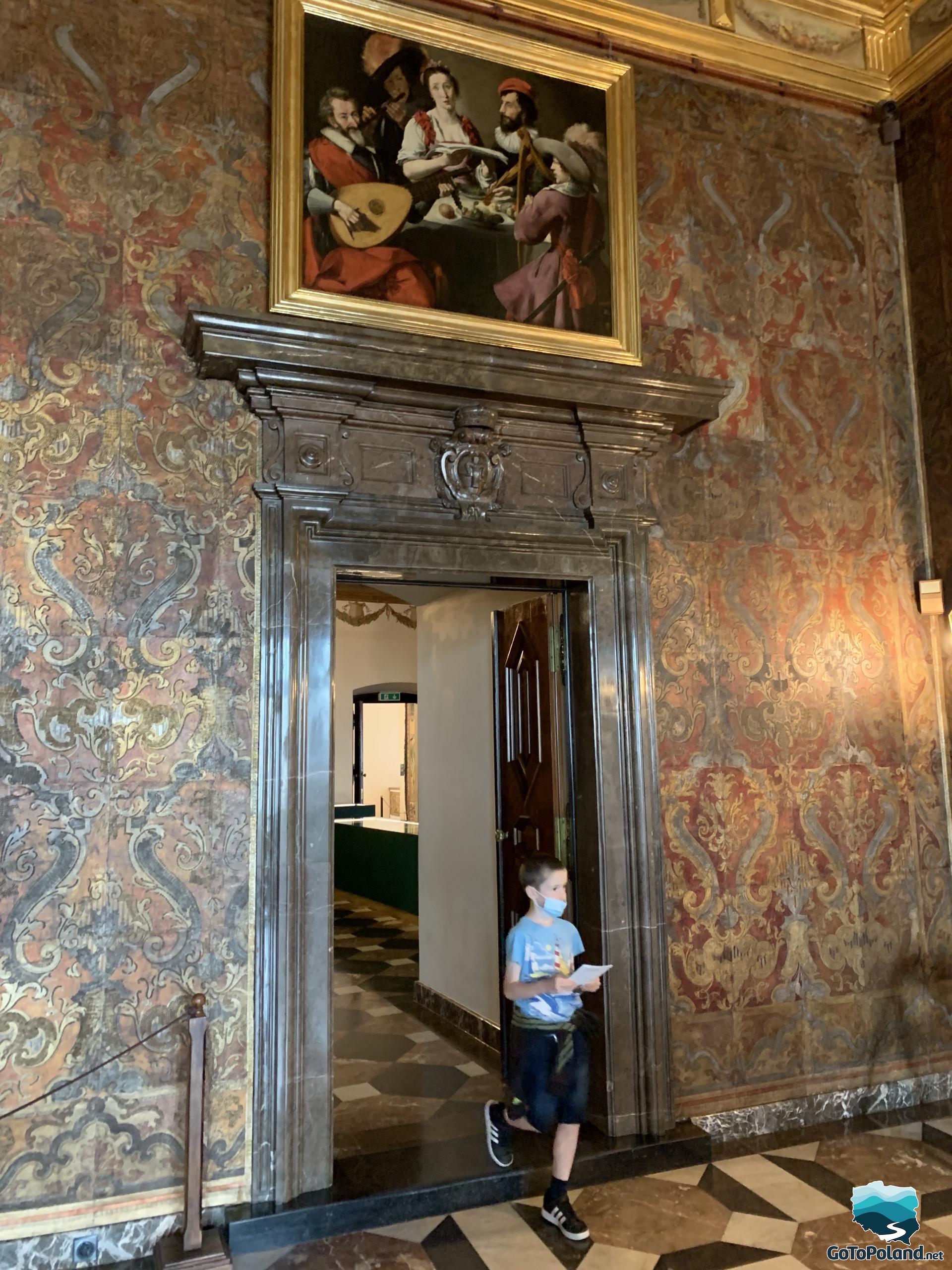 the boy enters the chamber through a large door, the walls are covered with fabrics, wallpaper or paintings, a large picture hangs above the door