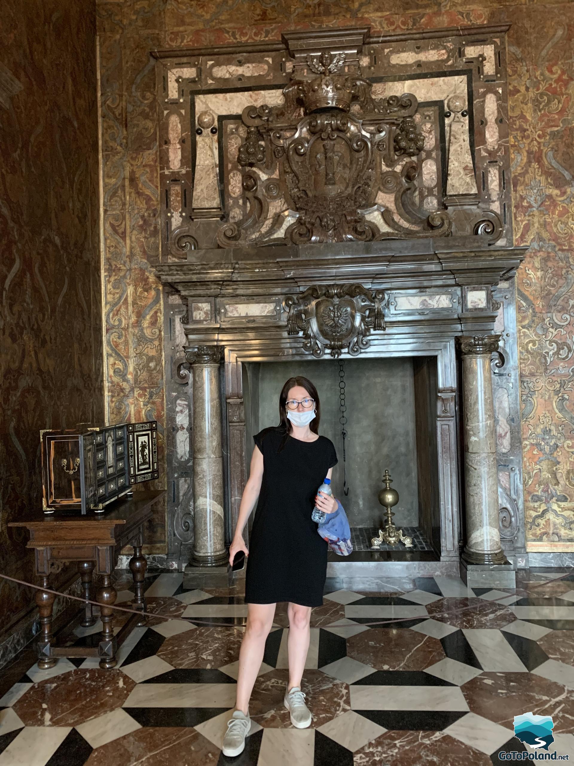 a woman is standing in a chamber in front of a decorative, large fireplace