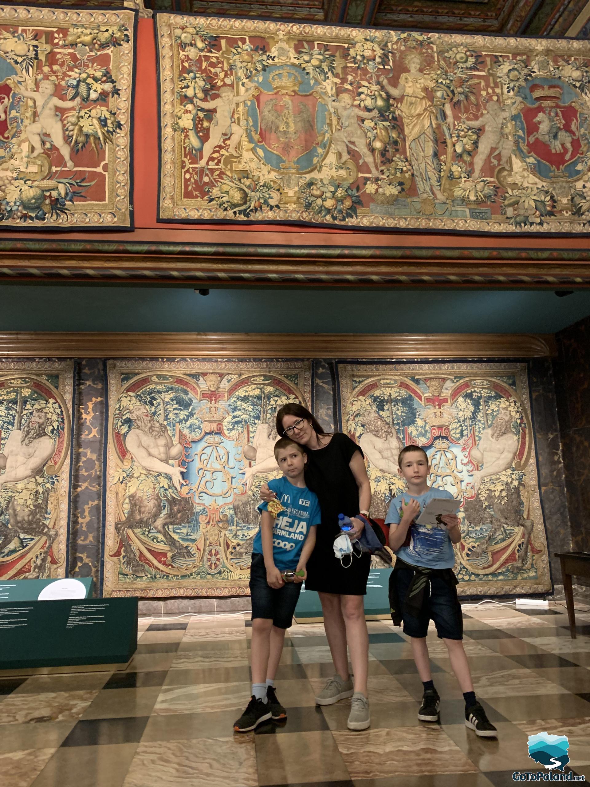 a woman and two boys are standing in a castle room, behind them hang two tapestries, one at the bottom and the other above