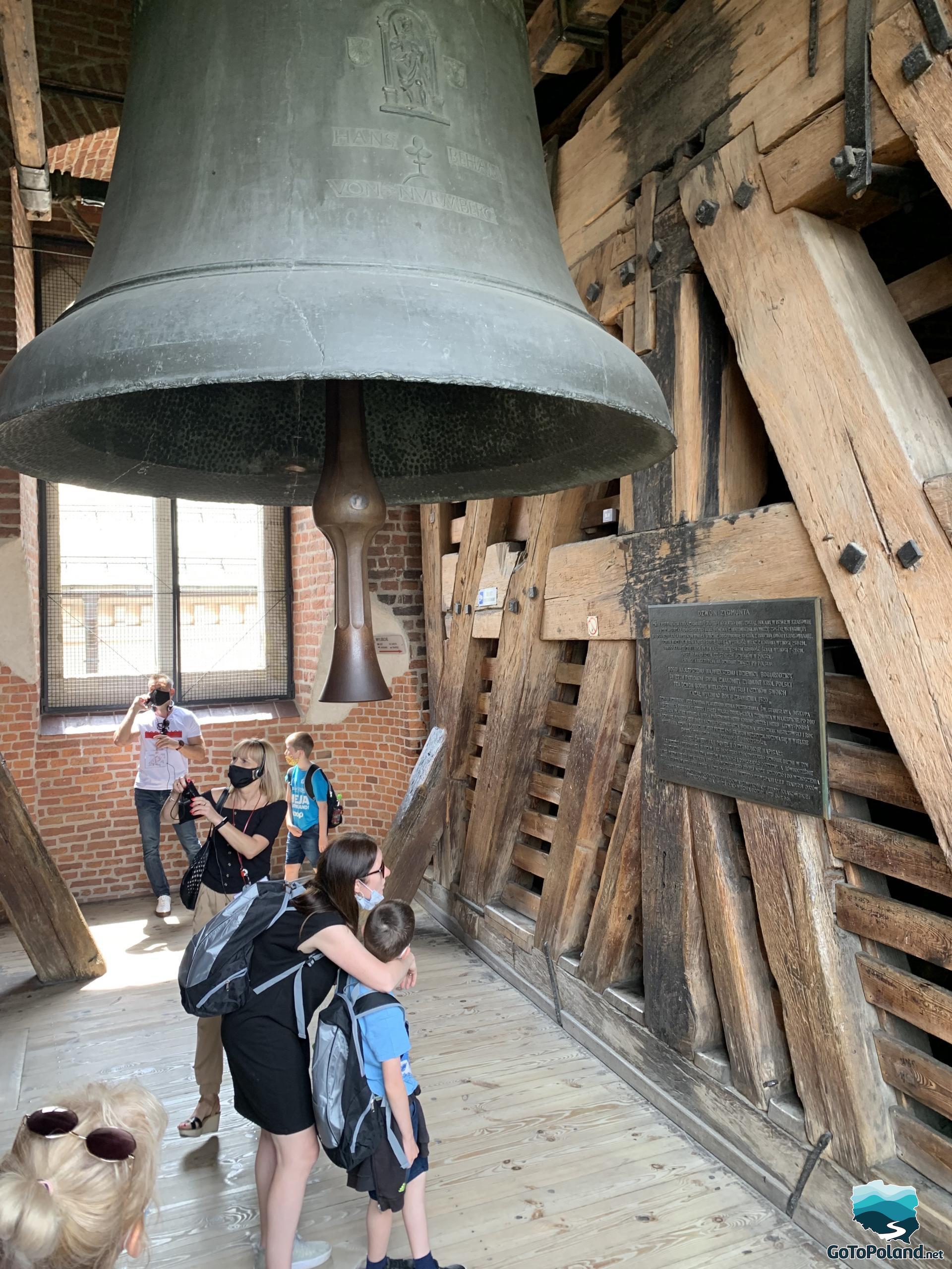 a woman an a boy are standing under a huge bell, she is reading an information about bell