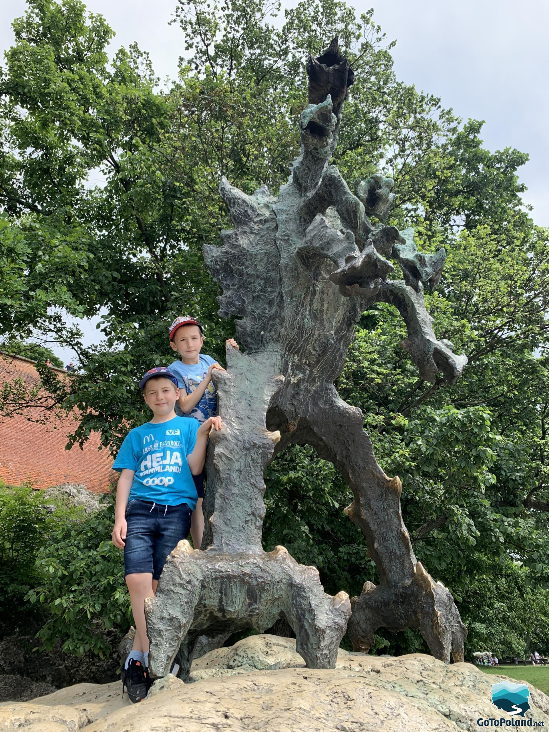two boys are standing by the dragon statue, trees behind them