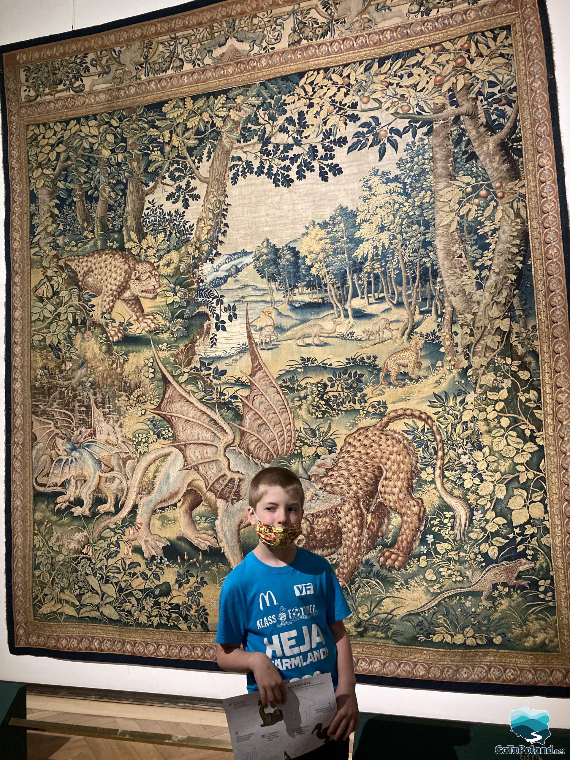a boy is standing in front of the tapestry, on the tapestry are tress and wild animals