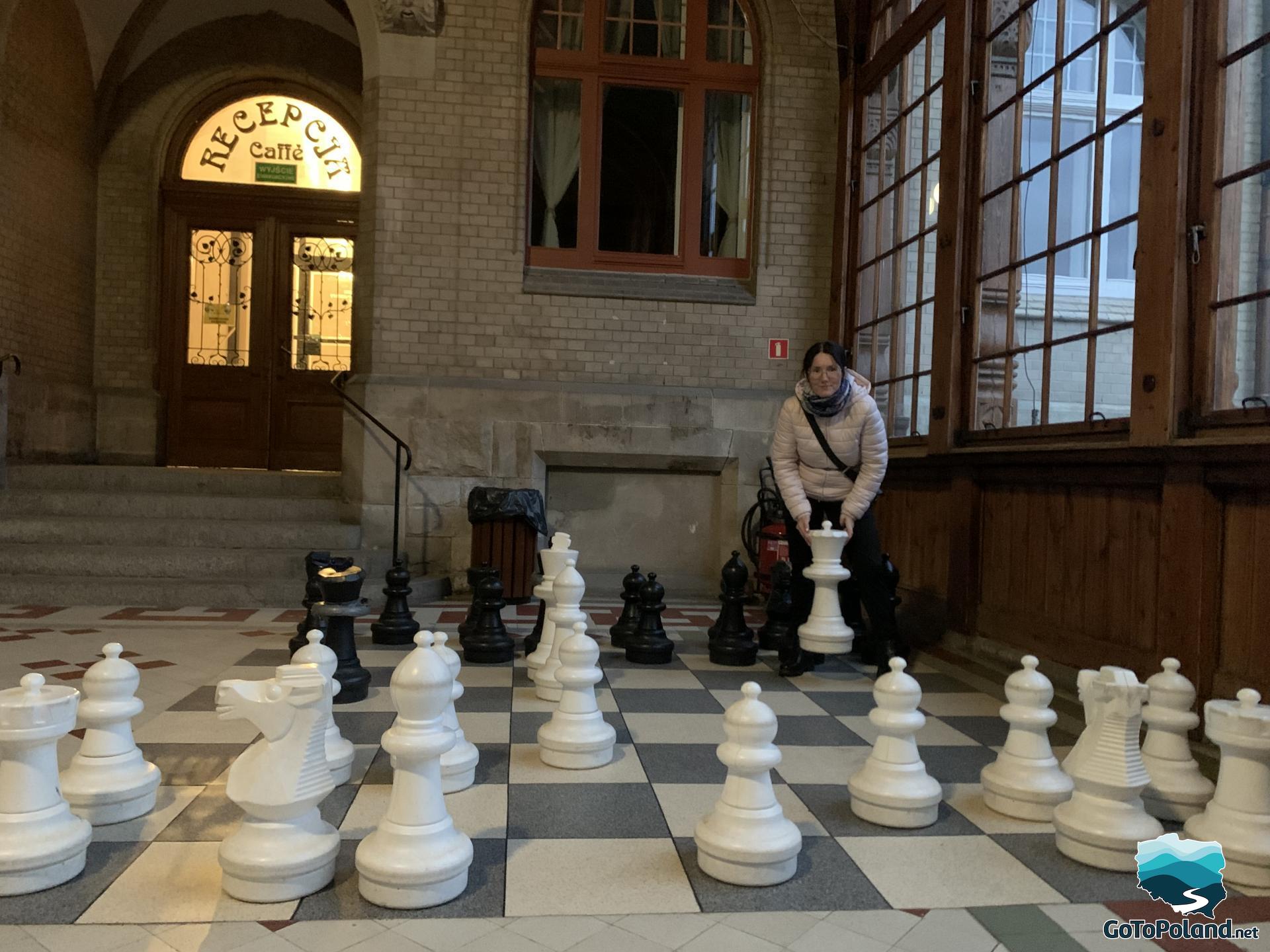 a woman is standing on a chessboard and picking up one white plastic chess piece