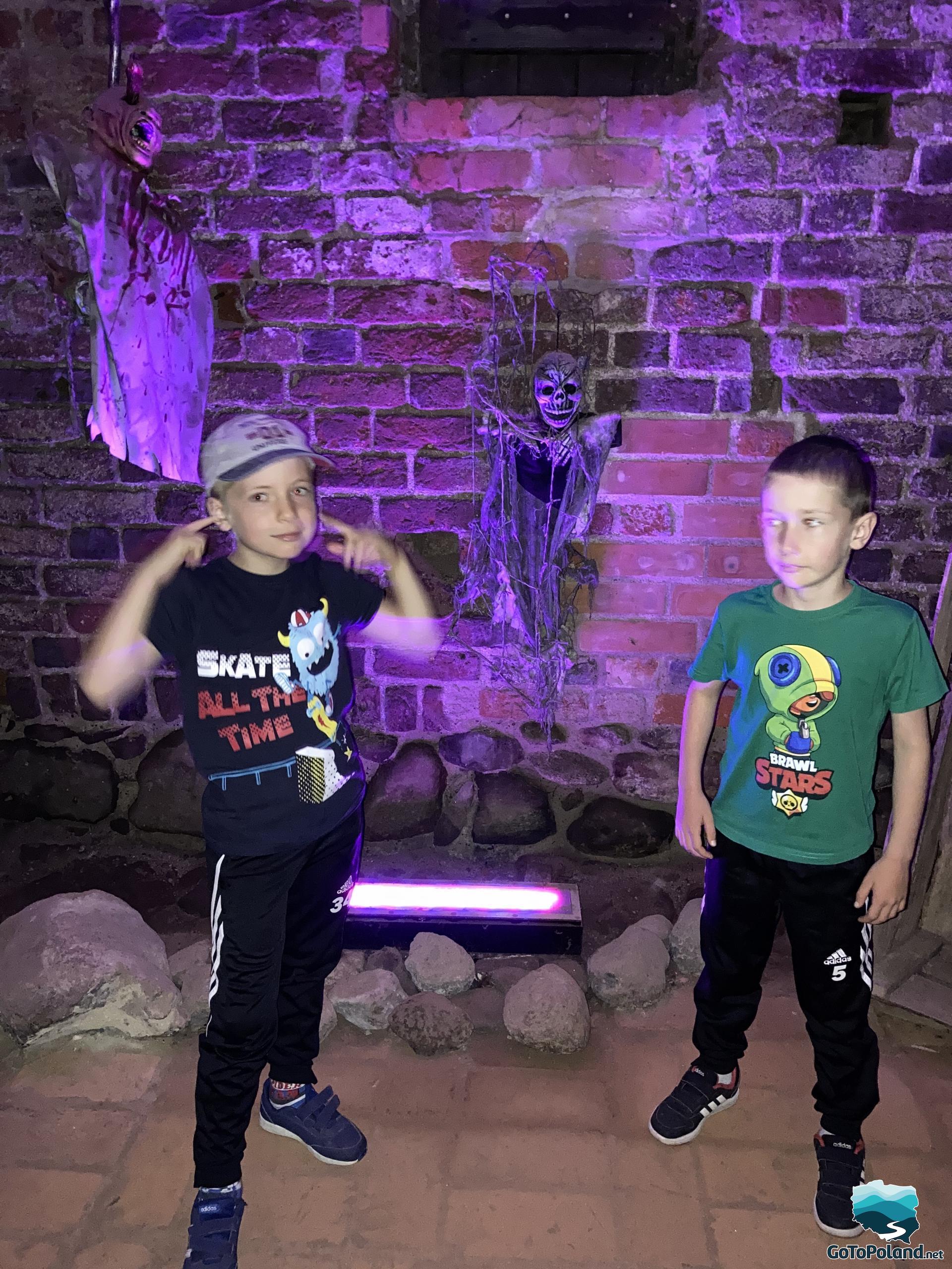 two children are standing in a room in a former castle, behind them a skeleton hangs on a wall, the light is purple
