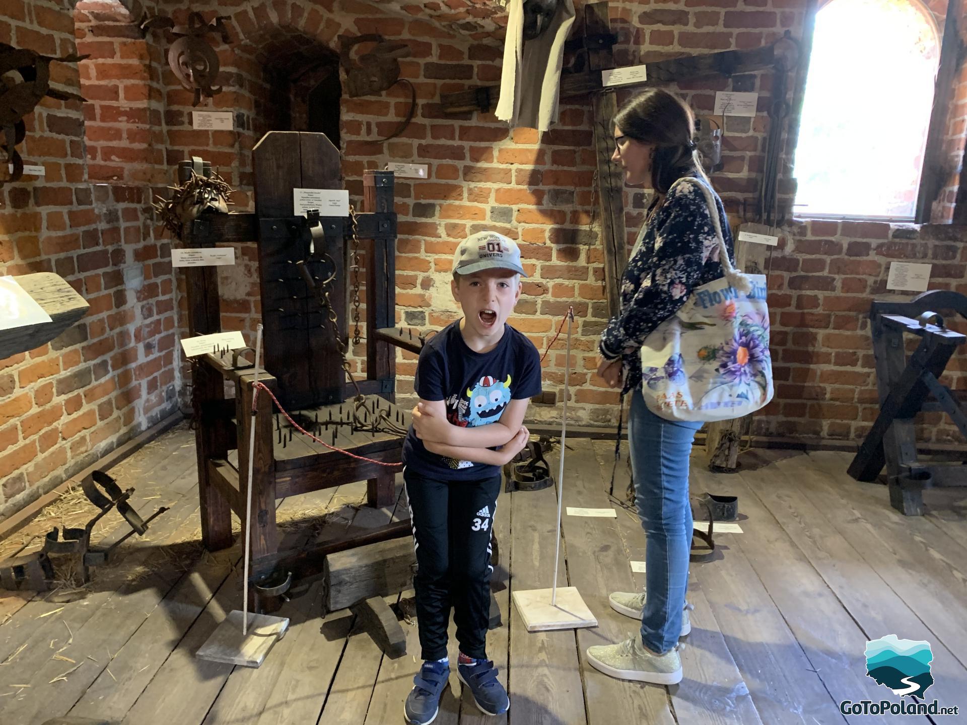 a woman and a boy are in a room which is filled with old instruments of torture, one of them is a chair with spikes