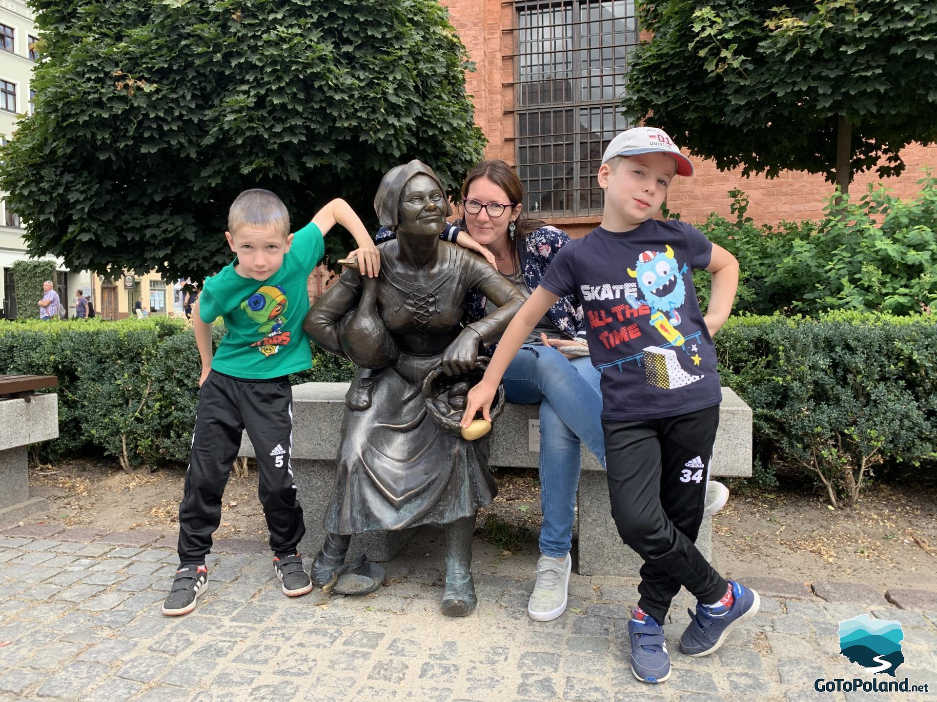 a bronze statue of a peasant woman holding a goose and a basket of eggs in the other hand, the statue of the woman is on a bench, next to her is a woman and two boys, one of them is touching a golden egg, which according to legend is supposed to bring good luck