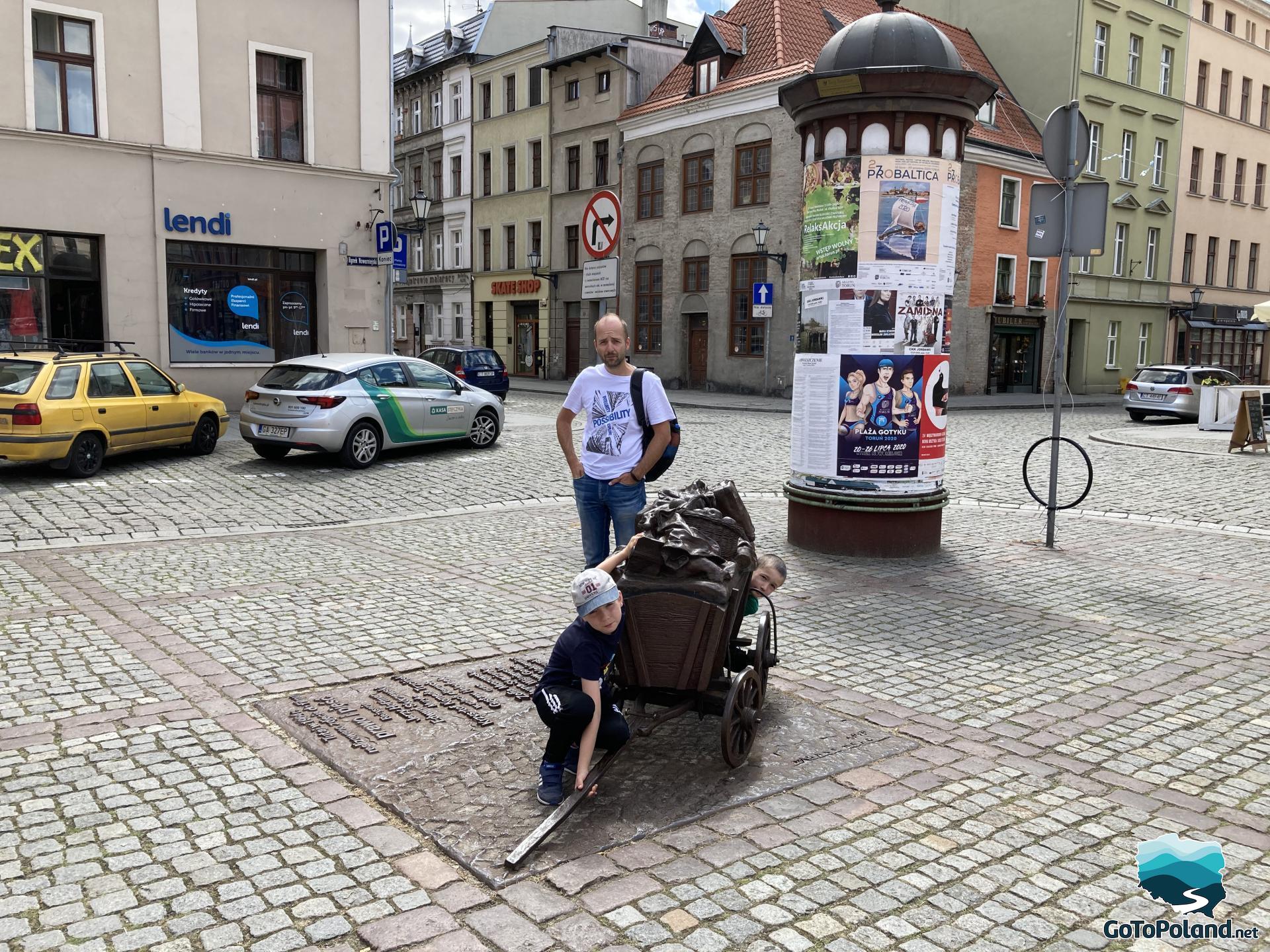 a man with two children are next to the wheelbarrow statue, there is a commemorative plaque from the movie The Law and the Fist, which was shot in this place