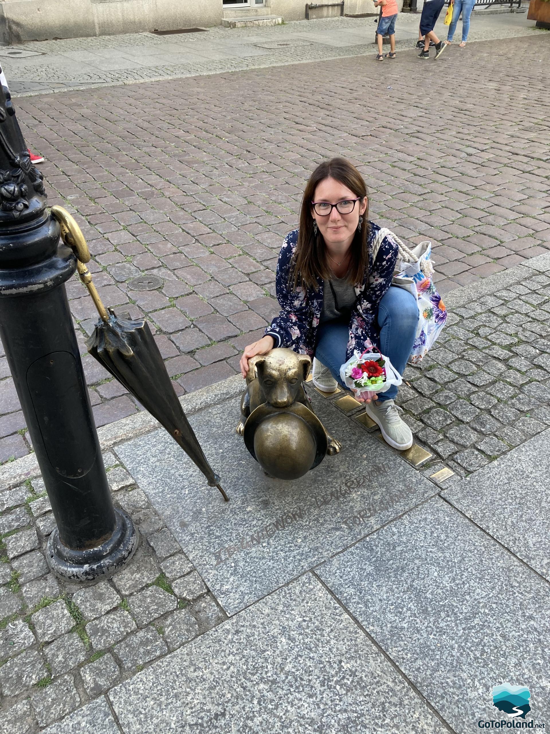 a woman is kneeling next to the dog statue, there is also un umbrella leaning on a street lamp