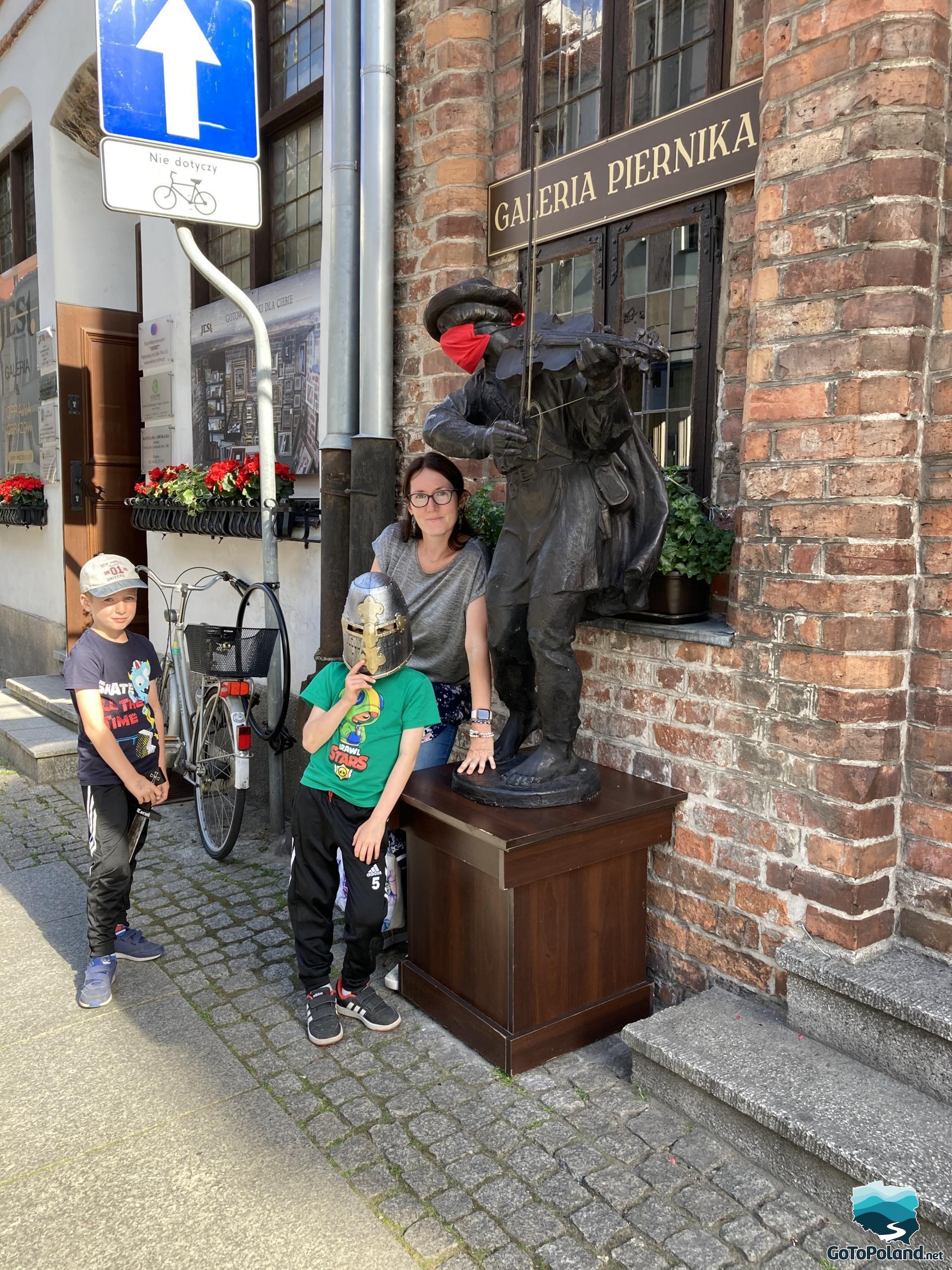 a woman and two children are standing near the statue of a violinist who has a red anti-covid mask and a violin in his hand, the figurine is in front of the Gingerbread Gallery