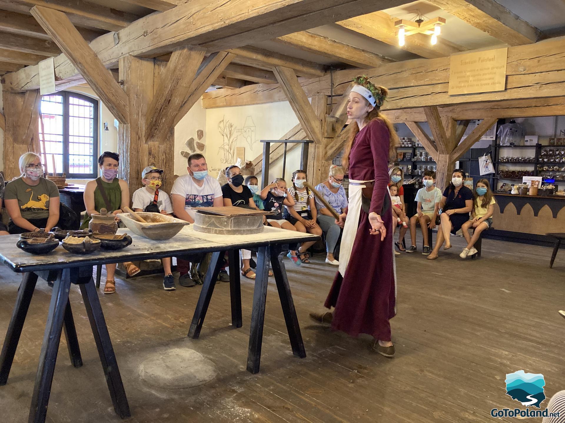 there are many tourists in the room who participate in gingerbread baking, the woman conducting the classes is wearing a flower wreath and a long burgundy dress