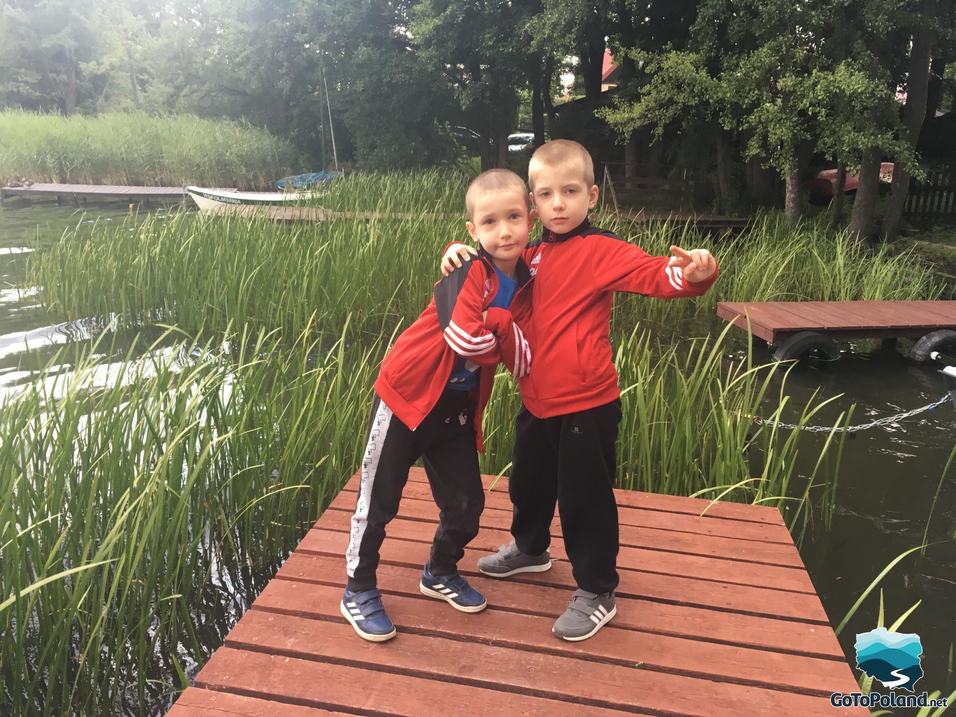 two boys are standing on a pier, there is a lake and more piers. Reeds grow in a lake
