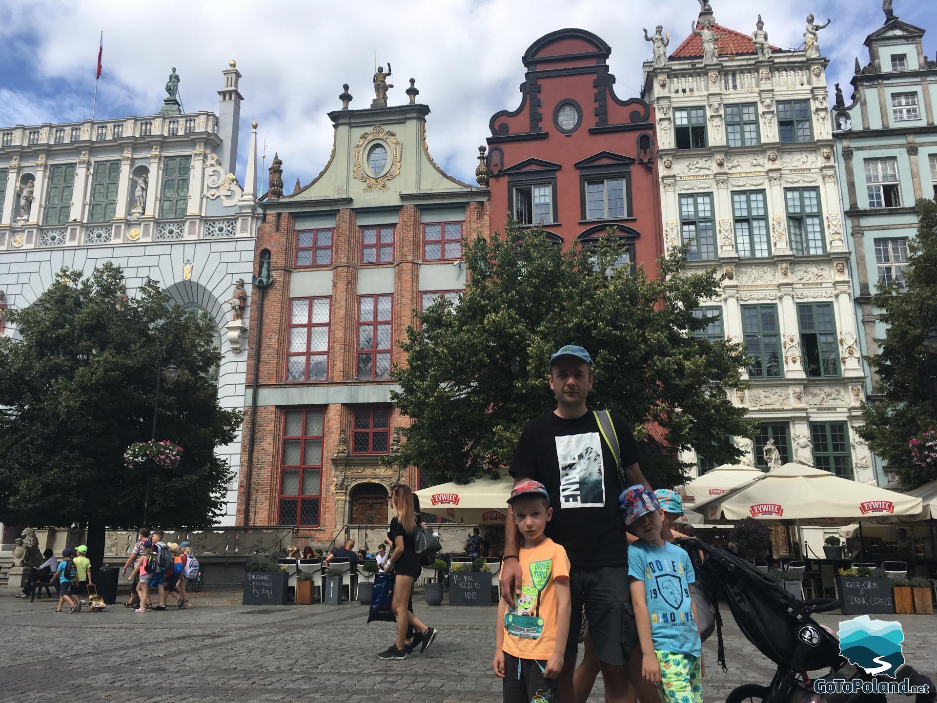 a man and two children are in the Old Market square, behind them are three trees and colourful tenement houses