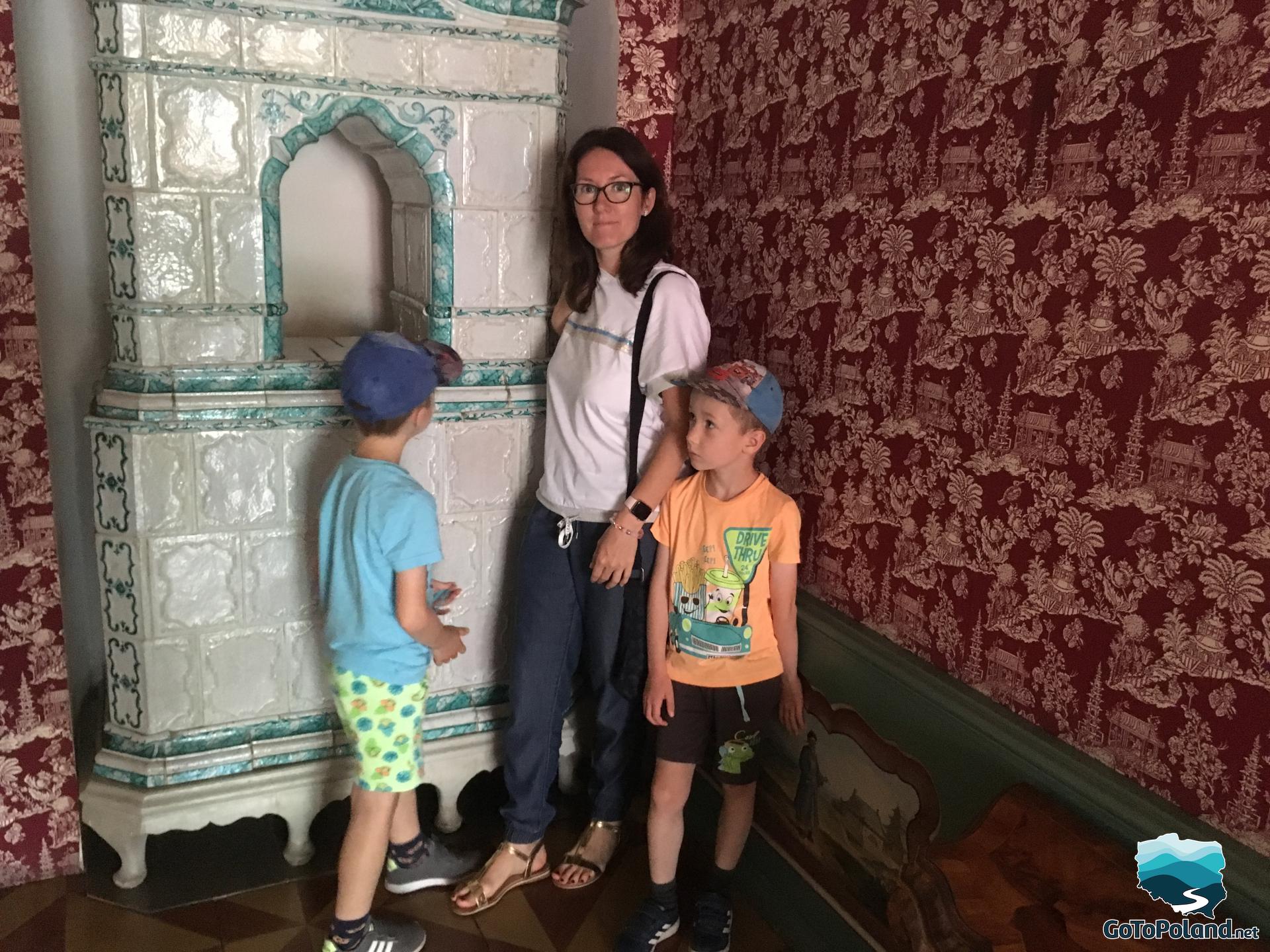 a woman and two boys standing near tiled stove, on the wall there is a patterned red wallpaper 