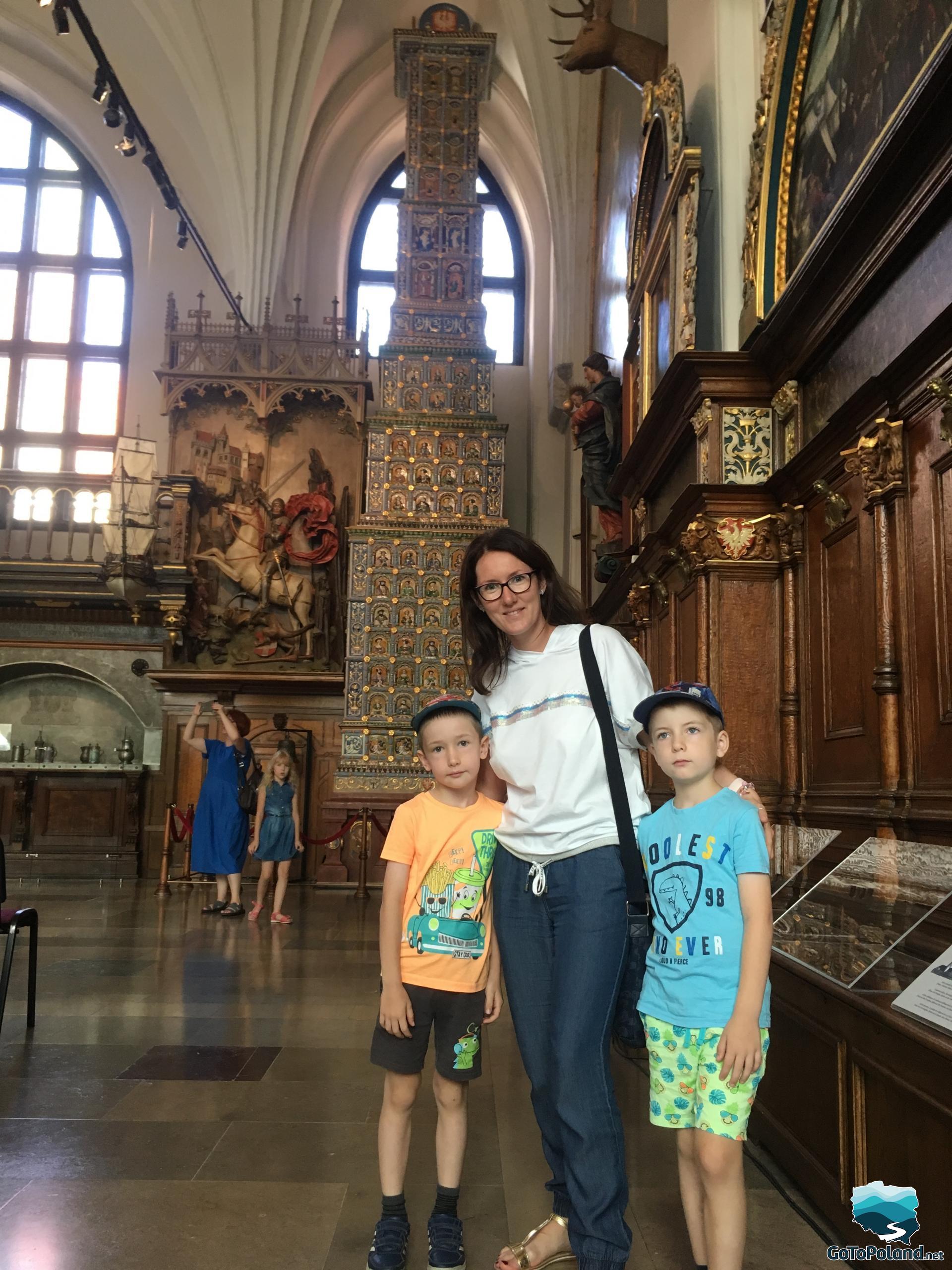 a woman and two boys are in a very decorative hall, there is a tiled stove behind them