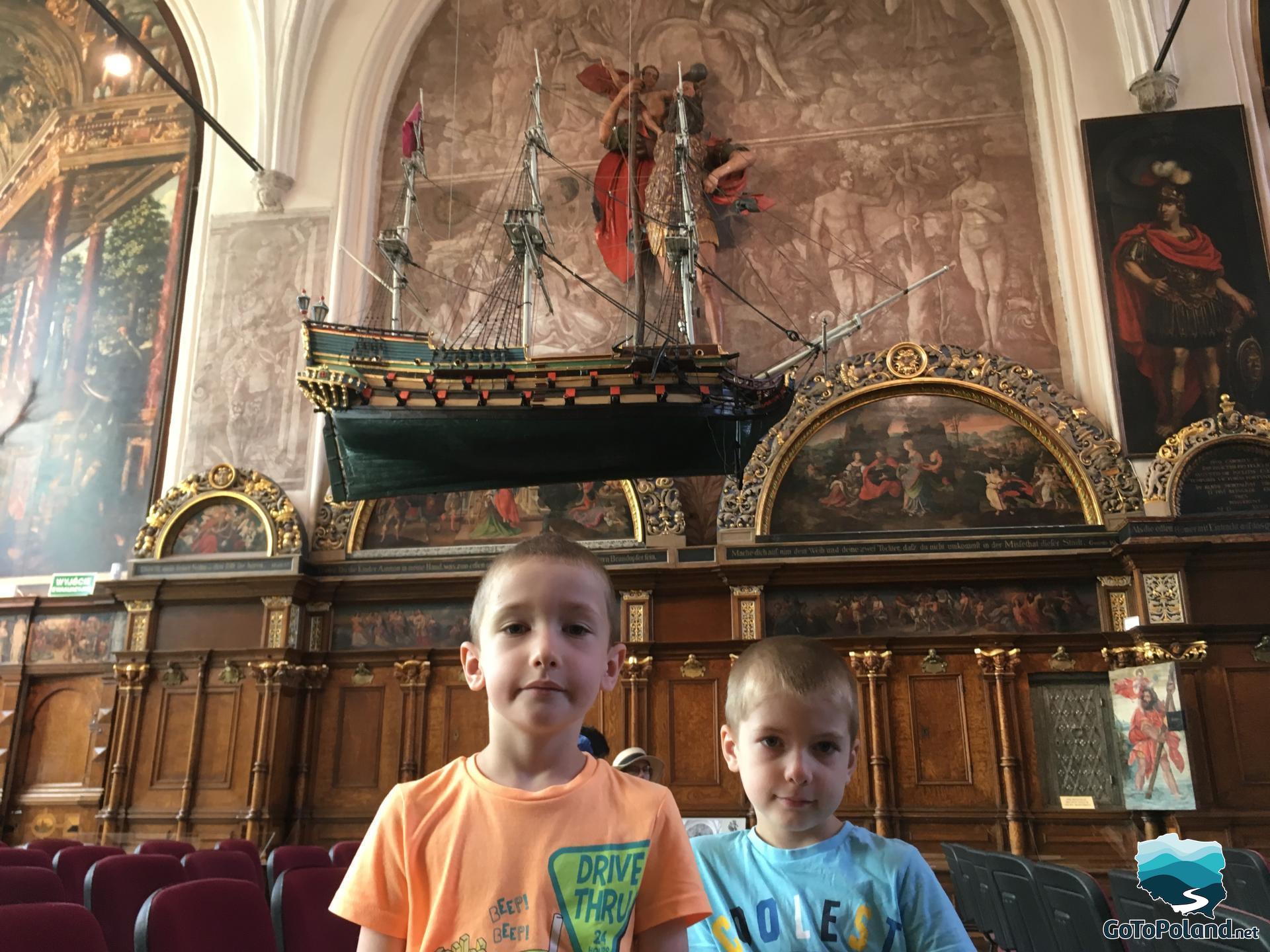 a two boys are in a hall, a model of a green ship hangs from the ceiling, walls are painted 
