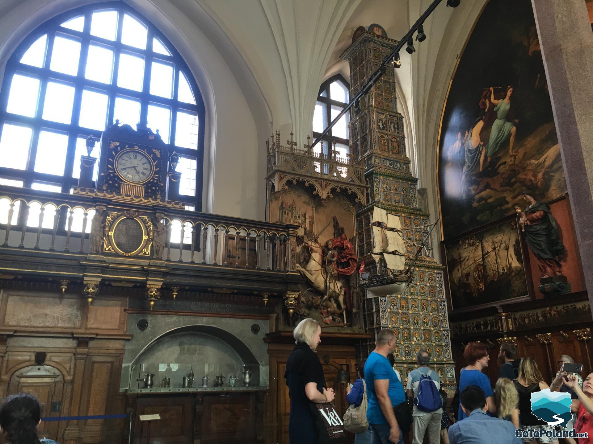 a large, decorative hall, an antique clock hanging on the wall, a huge tiled stove, many tourists