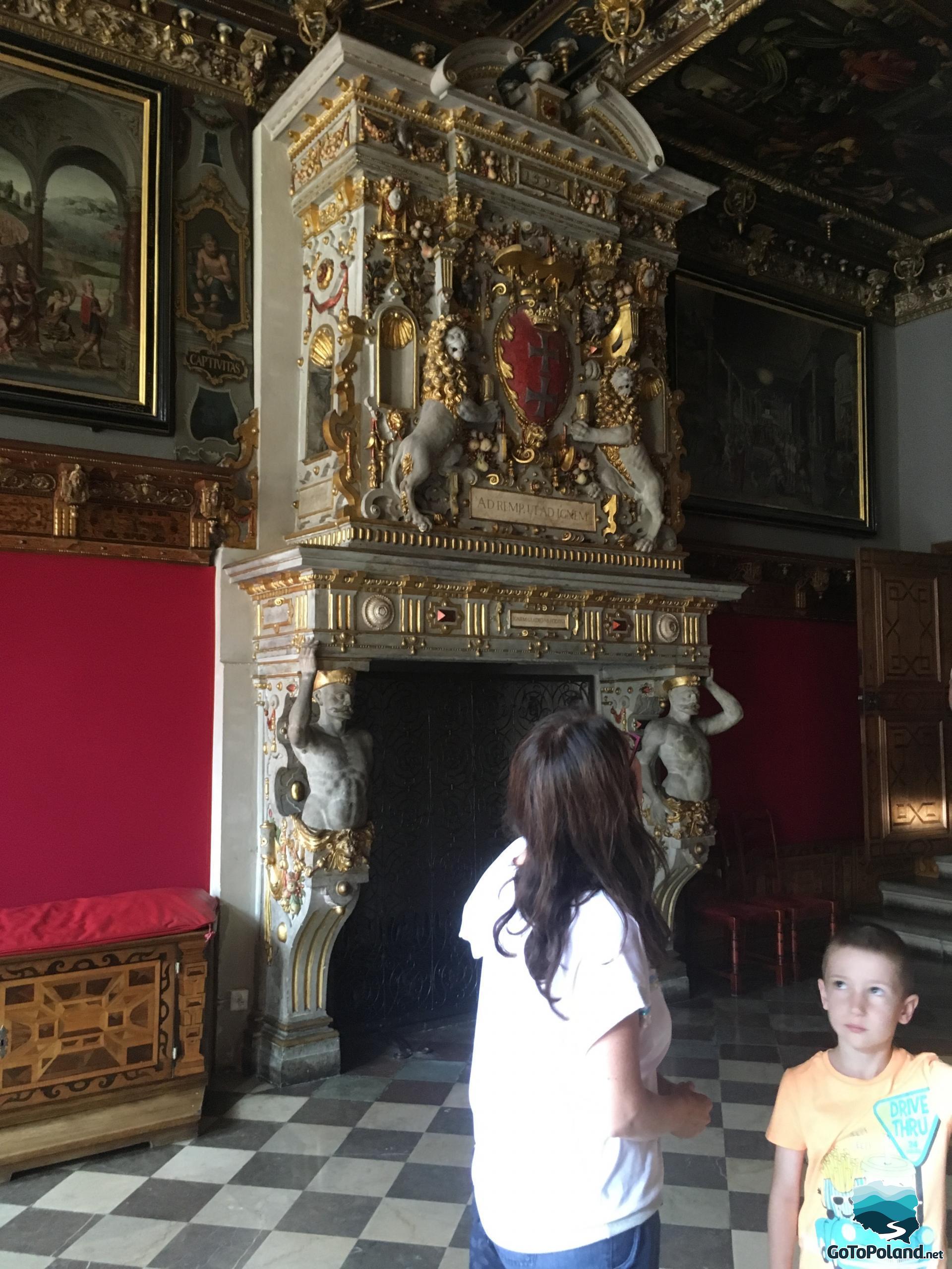 a woman and a boy standing in a decorative room, in from of a historic stove with gold ornaments