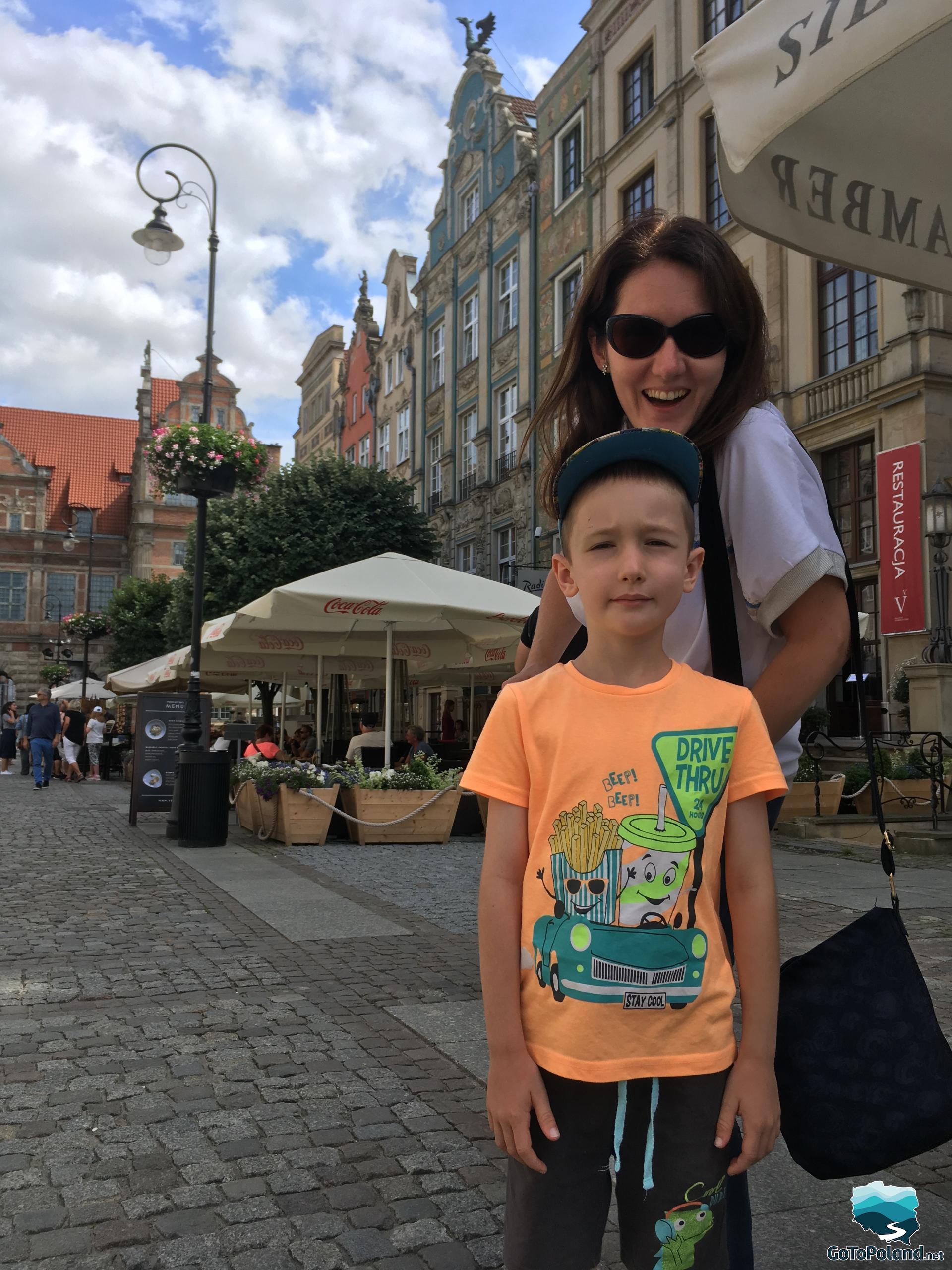 a woman and a boy are in a historic center, tenement houses around them