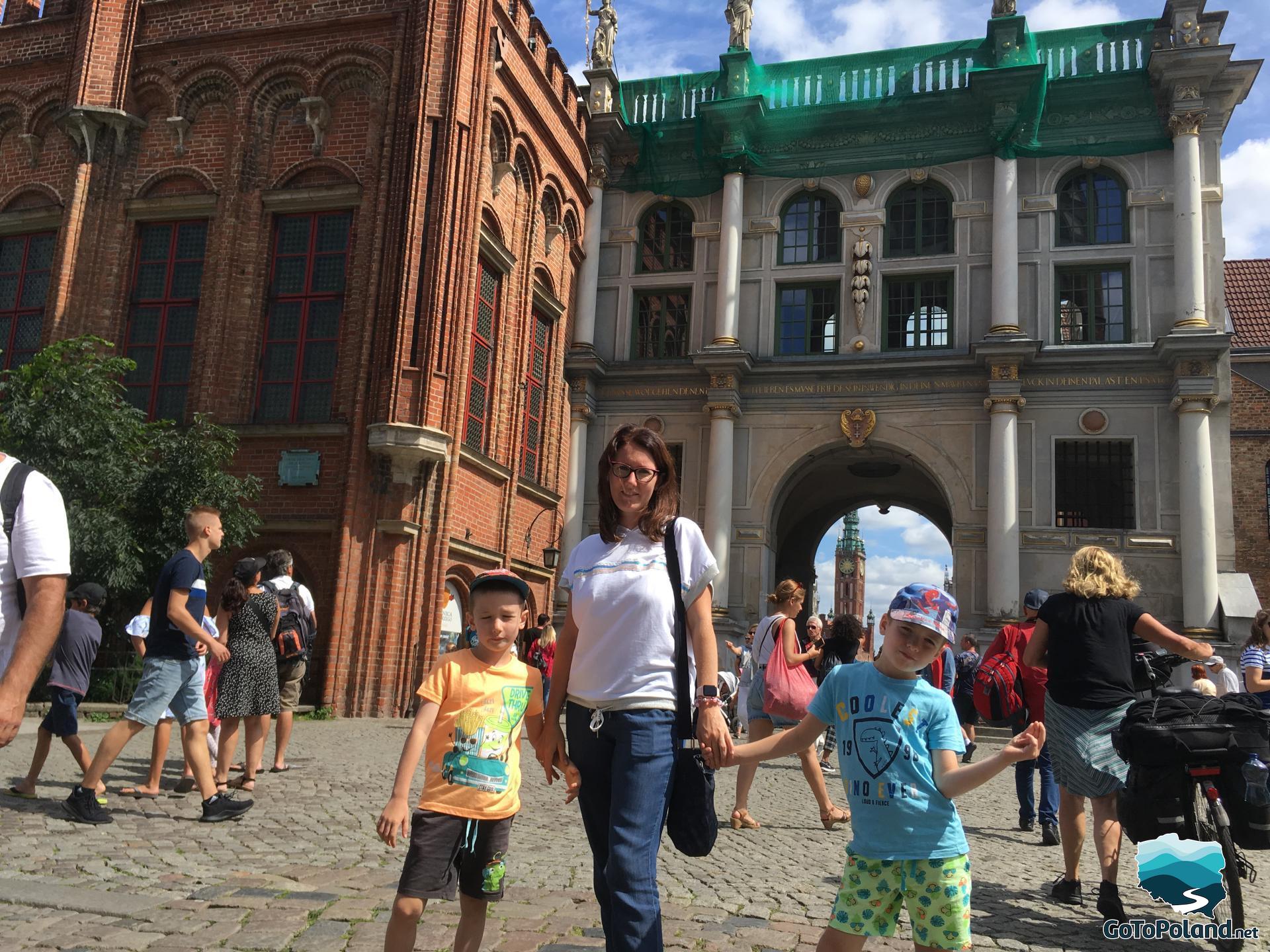 a woman with two children are standing by a red brick building, this building is next to a light colored gate with greek style columns, many tourists are around them