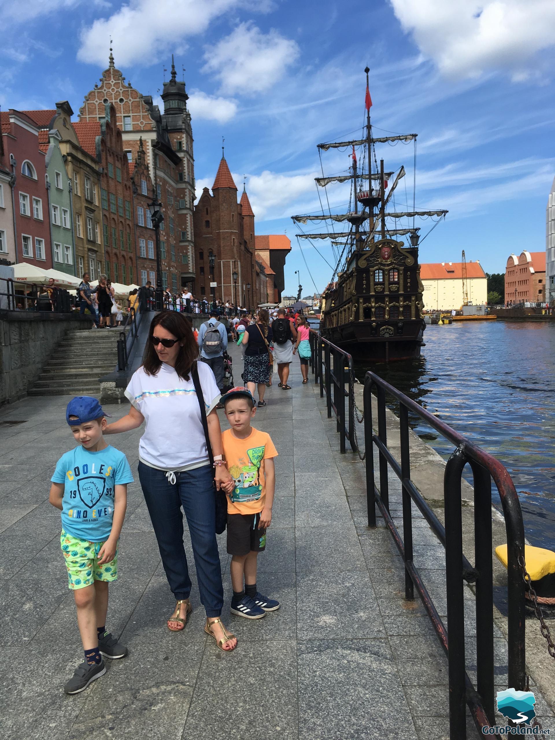 a woman and two children are standing by the wharf, an old crane can be seen in the background, a historic ship is standing on the river