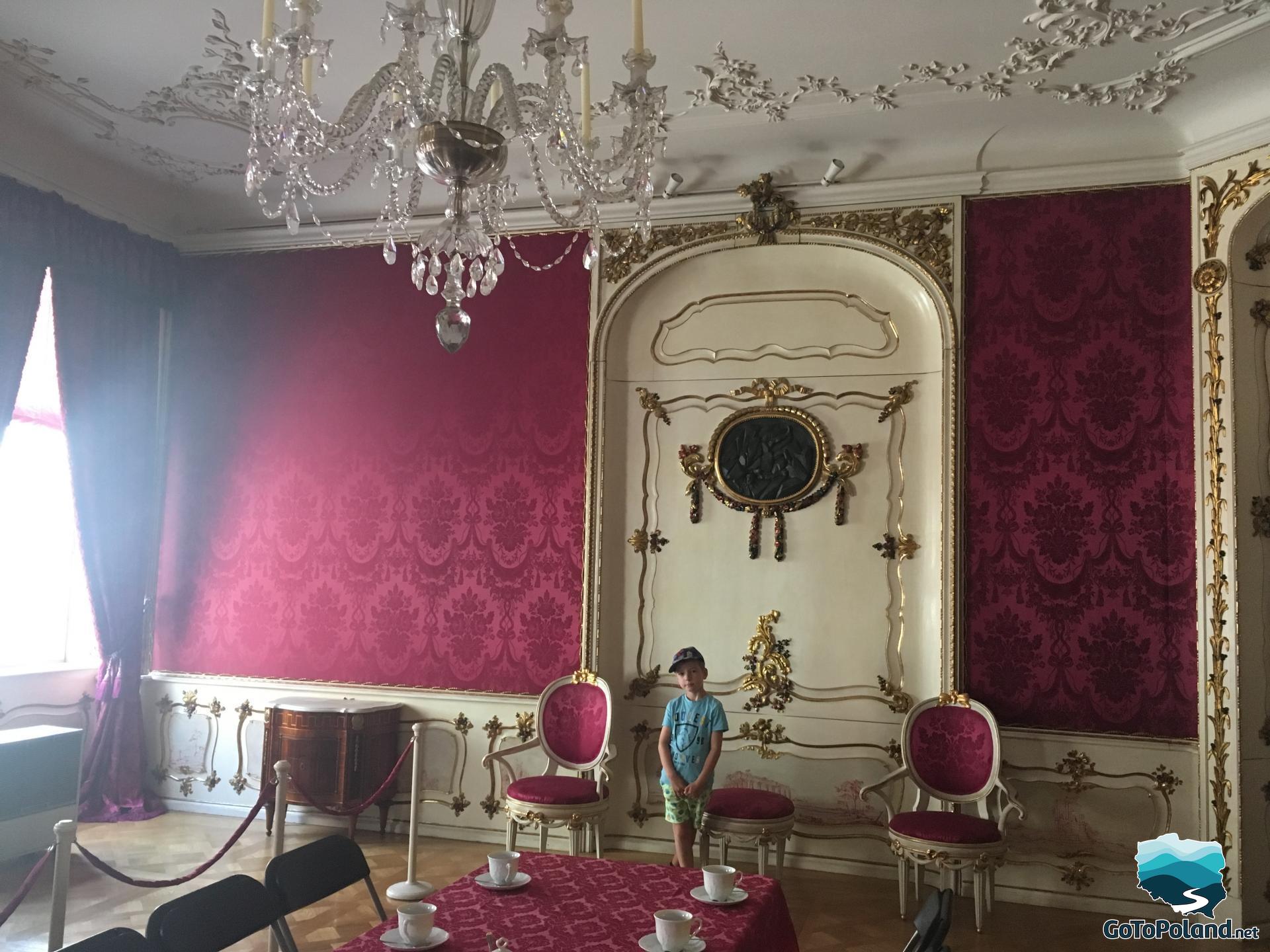 a boy is standing in a room, there is maroon fabric or wallpaper on the walls, a crystal chandelier hangs, there is also a table covered with a red tablecloth and chairs with upholstery in the same color