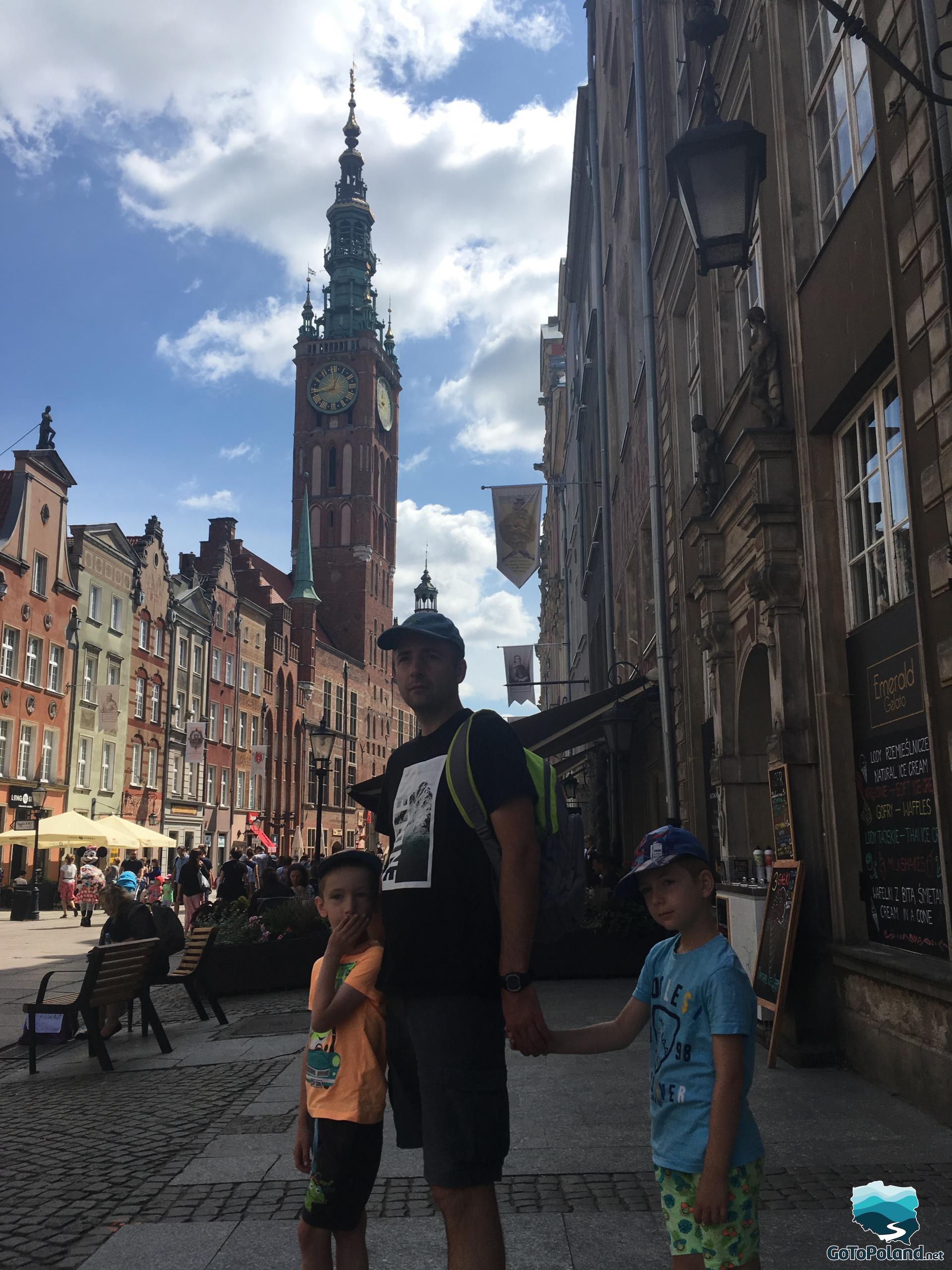 a man with two boys is standing in the street of the market square. In the background there is a historic Town Hall with beautiful, high tower, around them there are tenement houses and lots of tourists 