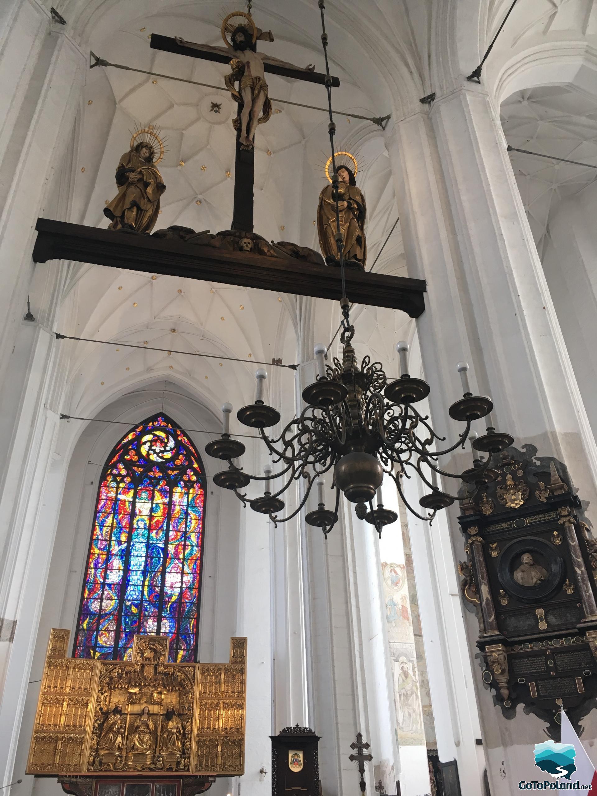 the interior of the basilica with a large colored stained glass window, a black chandelier, a golden triptych, the walls of the basilica are white, a cross with the crucified Christ hangs from the ceiling