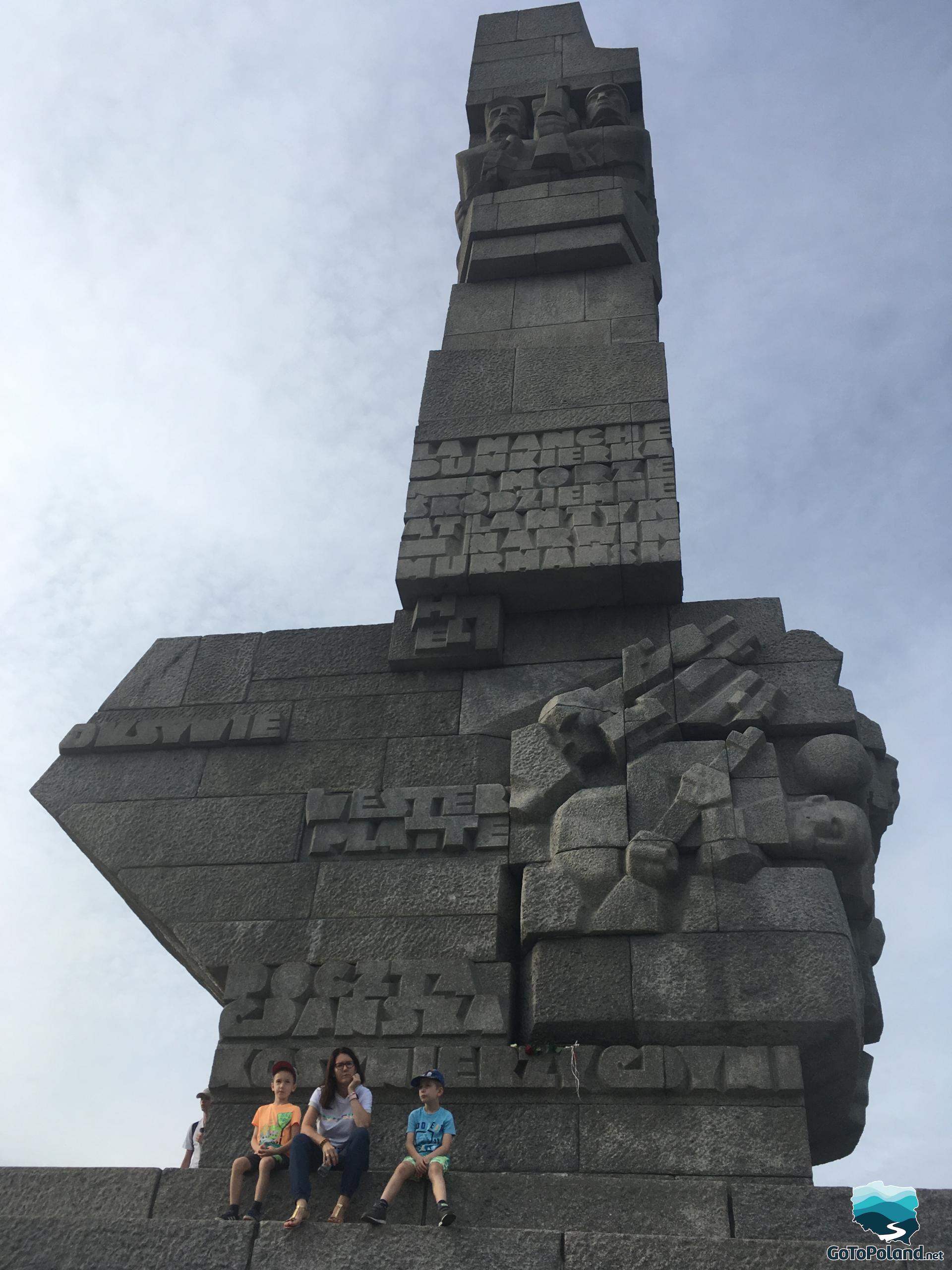 a woman and two kids sitting at the feet of a huge monument commemorating the soldiers who died defending Westerplatte