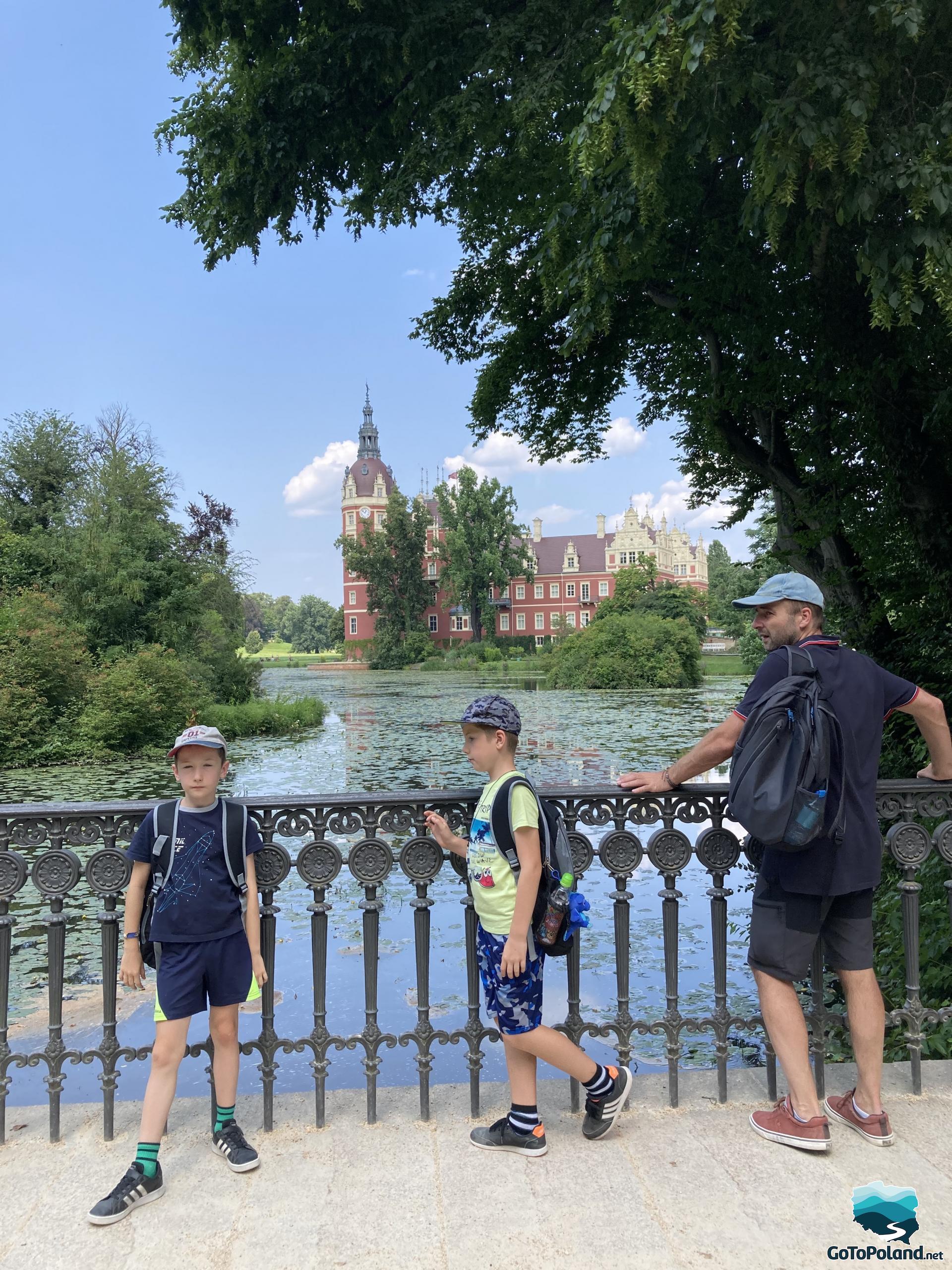 two boys and a man are standing on a small bridge, a pond and the castle are in the background