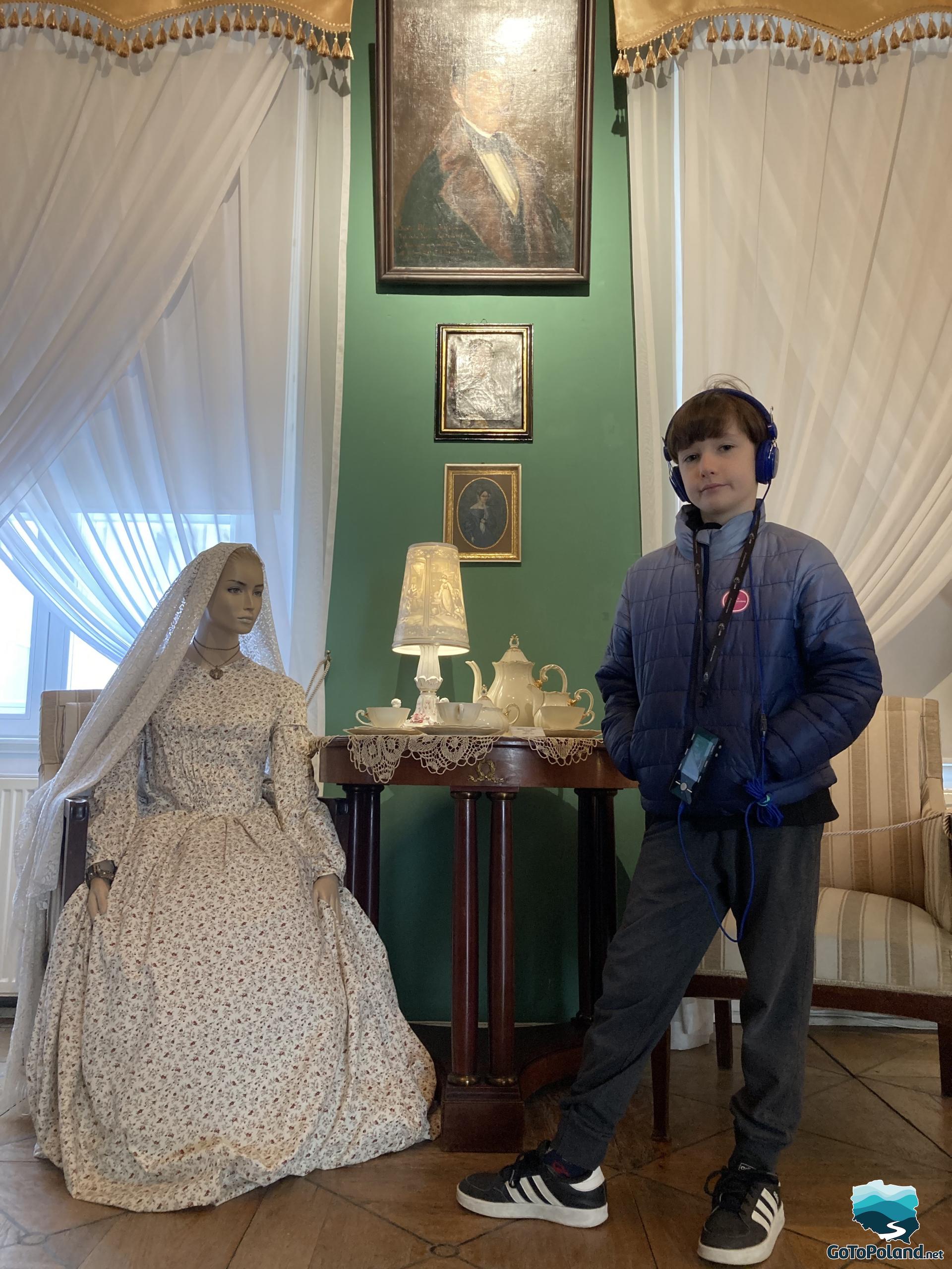 a boy is standing next to the artificial model of a woman, on the table there is a porcelain set